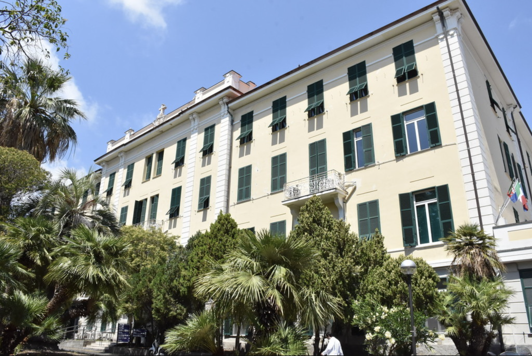 A large, pale yellow multi-story building with green shutters and small balconies, surrounded by lush tropical trees and bushes, under a bright blue sky.