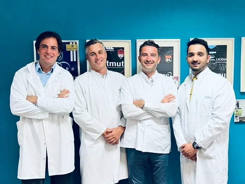 Four men in white lab coats standing in front of framed posters on a blue wall, smiling with arms crossed.