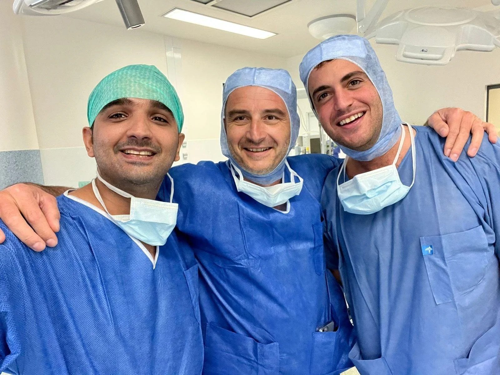 Three male medical professionals wearing blue scrubs, surgical masks, and caps, smiling and posing together in a hospital operating room.