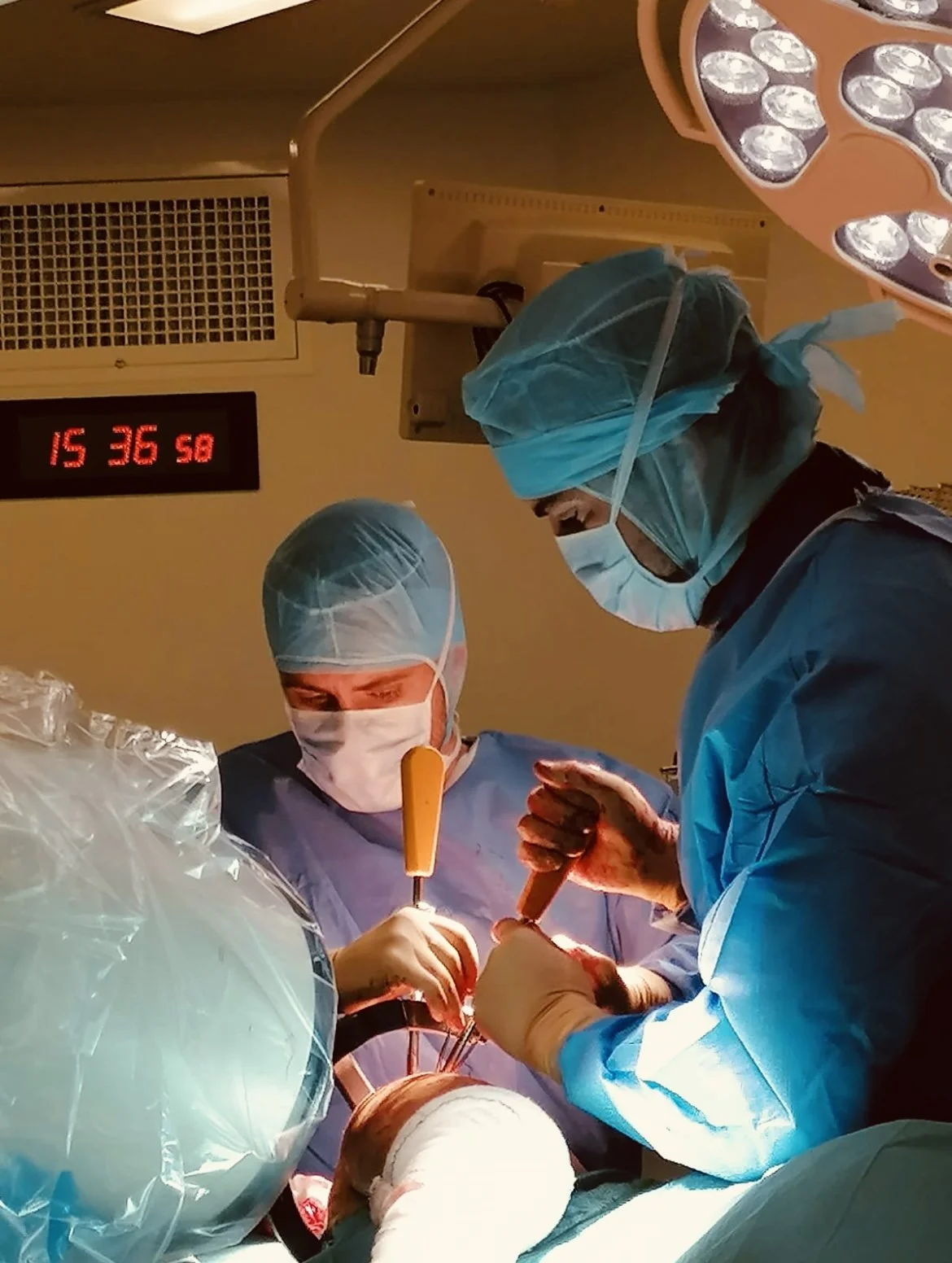 Two surgeons performing a procedure in an operating room, wearing surgical masks, gowns, and caps, with bright surgical lights overhead.
