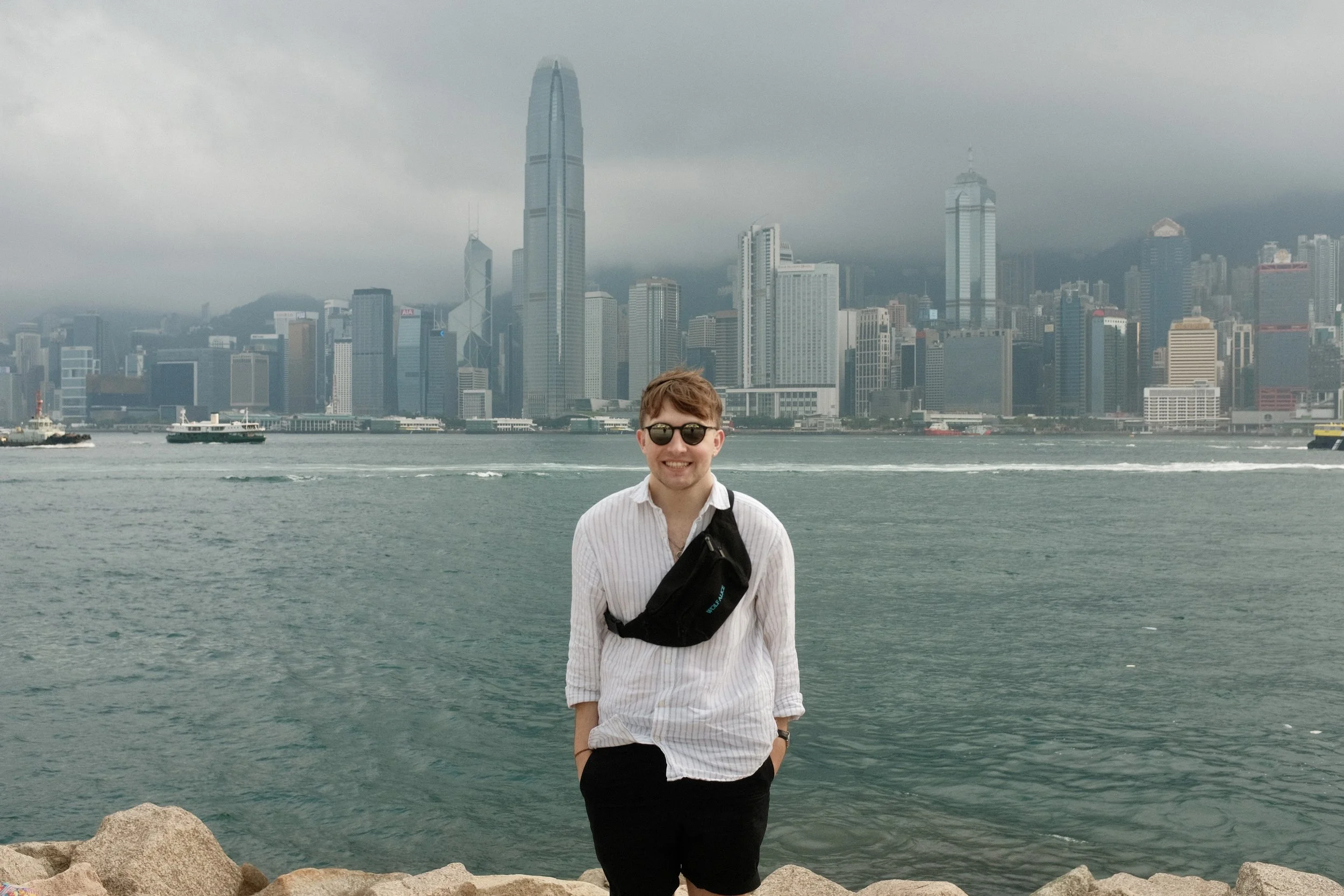A smiling young man wearing sunglasses, a white striped shirt, black shorts, and a black crossbody bag standing on rocks by the water with a city skyline and cloudy sky in the background.