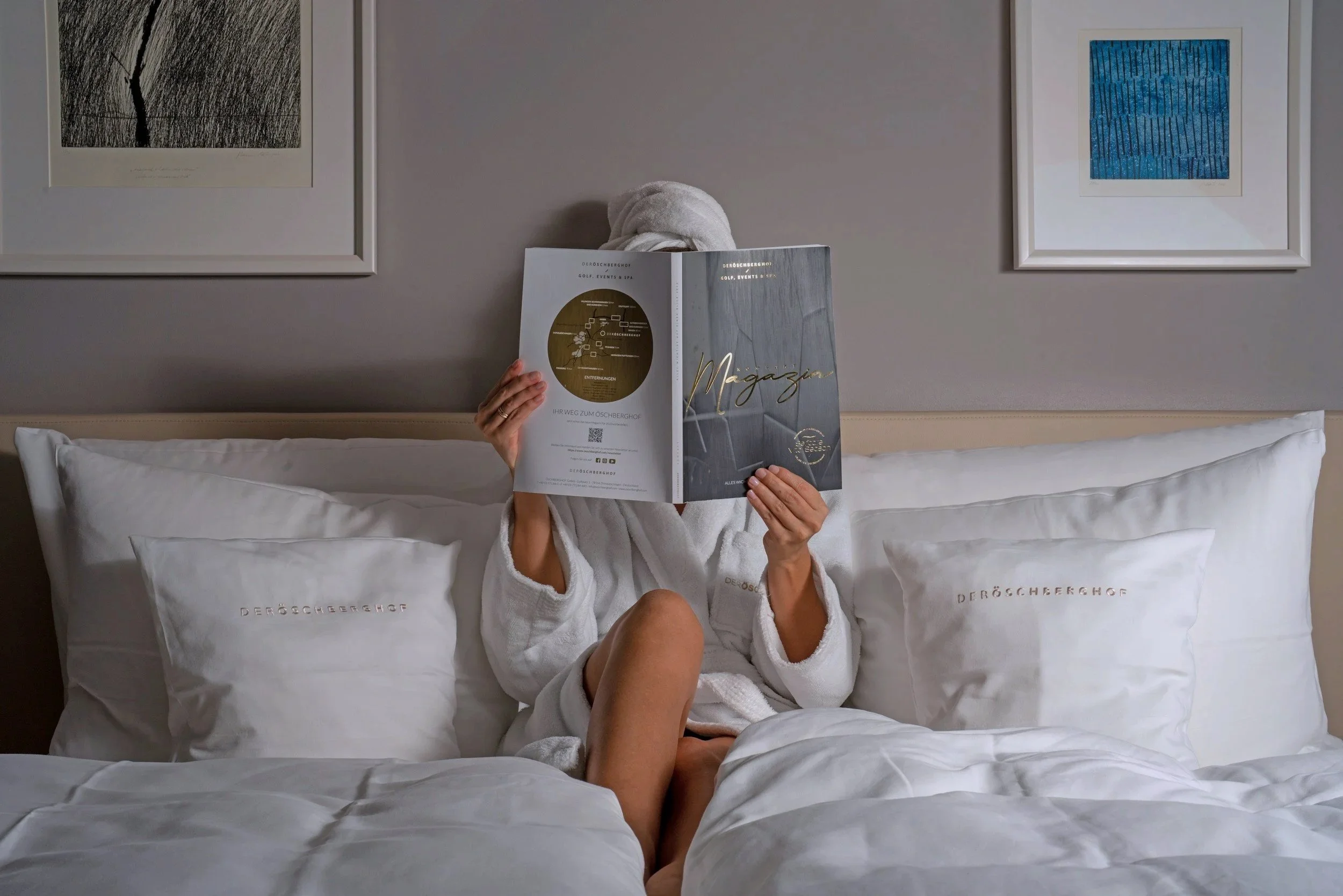 Person sitting on a bed, wearing a white robe, reading a magazine, with their face covered by it. The bed has white pillows and framed artwork hangs on the beige wall behind.