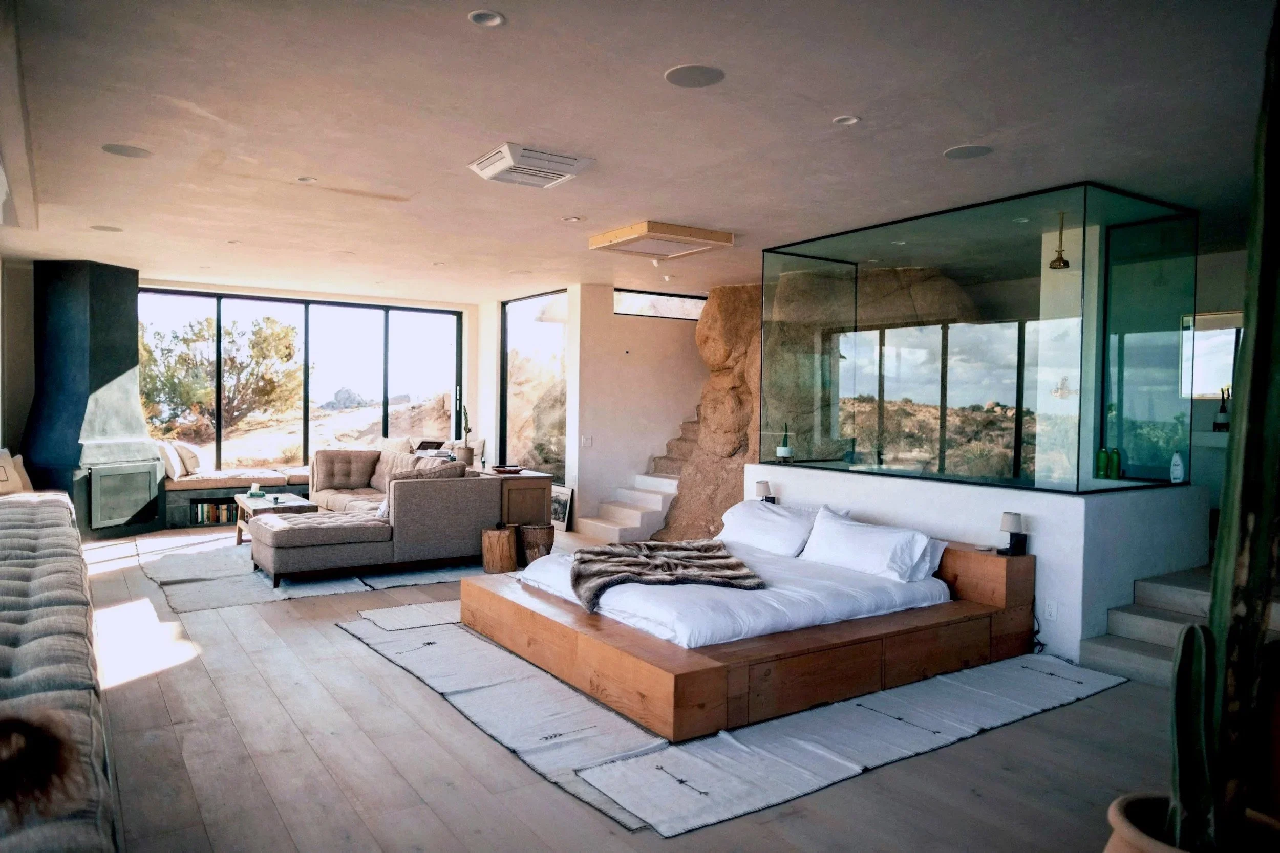 Modern luxury hotel suite living room with large curved beige sofa, assorted pillows, marble and black side tables, a flat-screen TV, art on the wall, and floor-to-ceiling windows showing a city skyline at sunset.