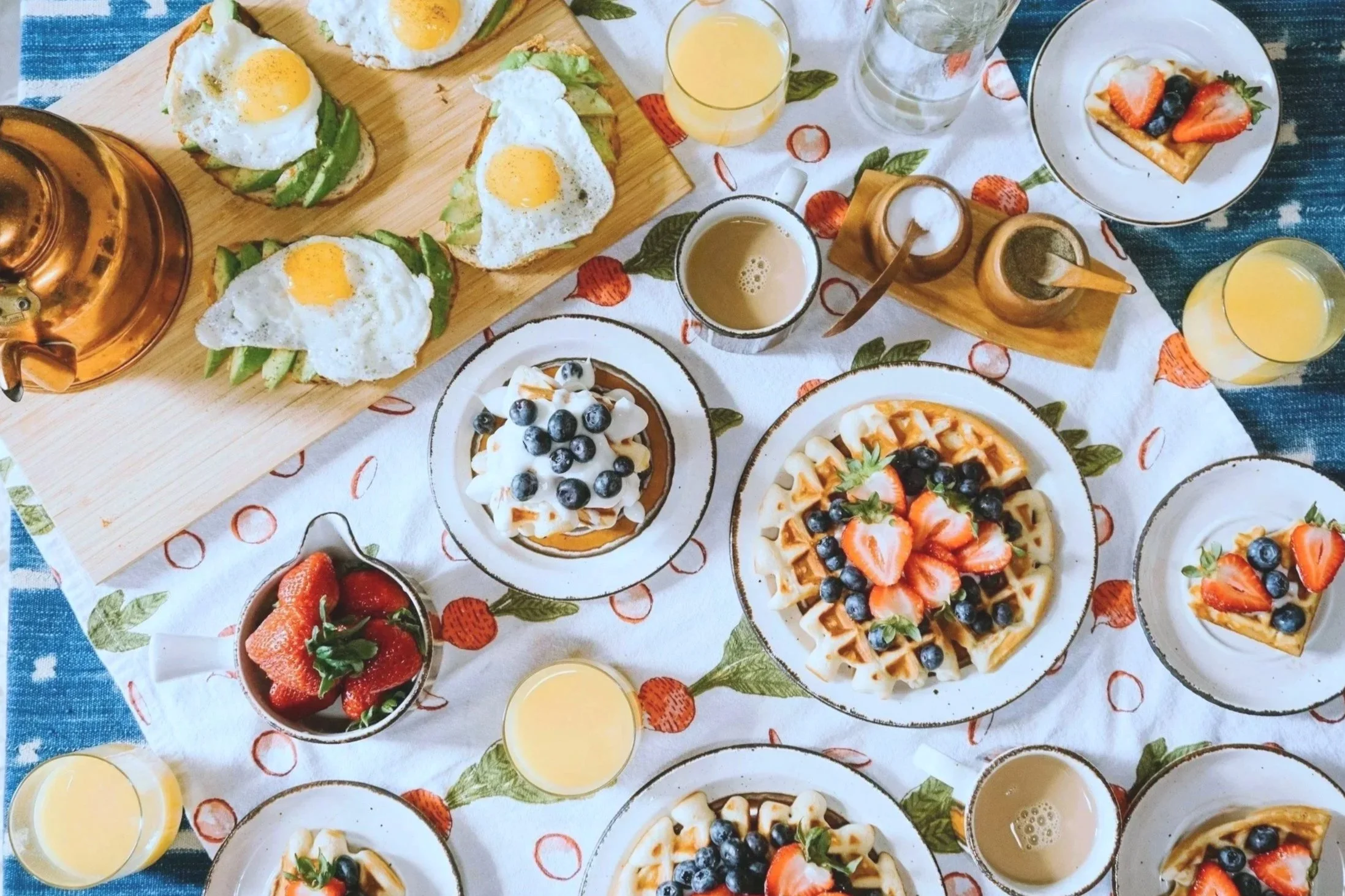 Breakfast spread featuring waffles topped with strawberries and blueberries, avocado toast with fried eggs, bowls of strawberries, glasses of orange juice, coffee, and syrup on a table with a white and strawberry-patterned tablecloth.