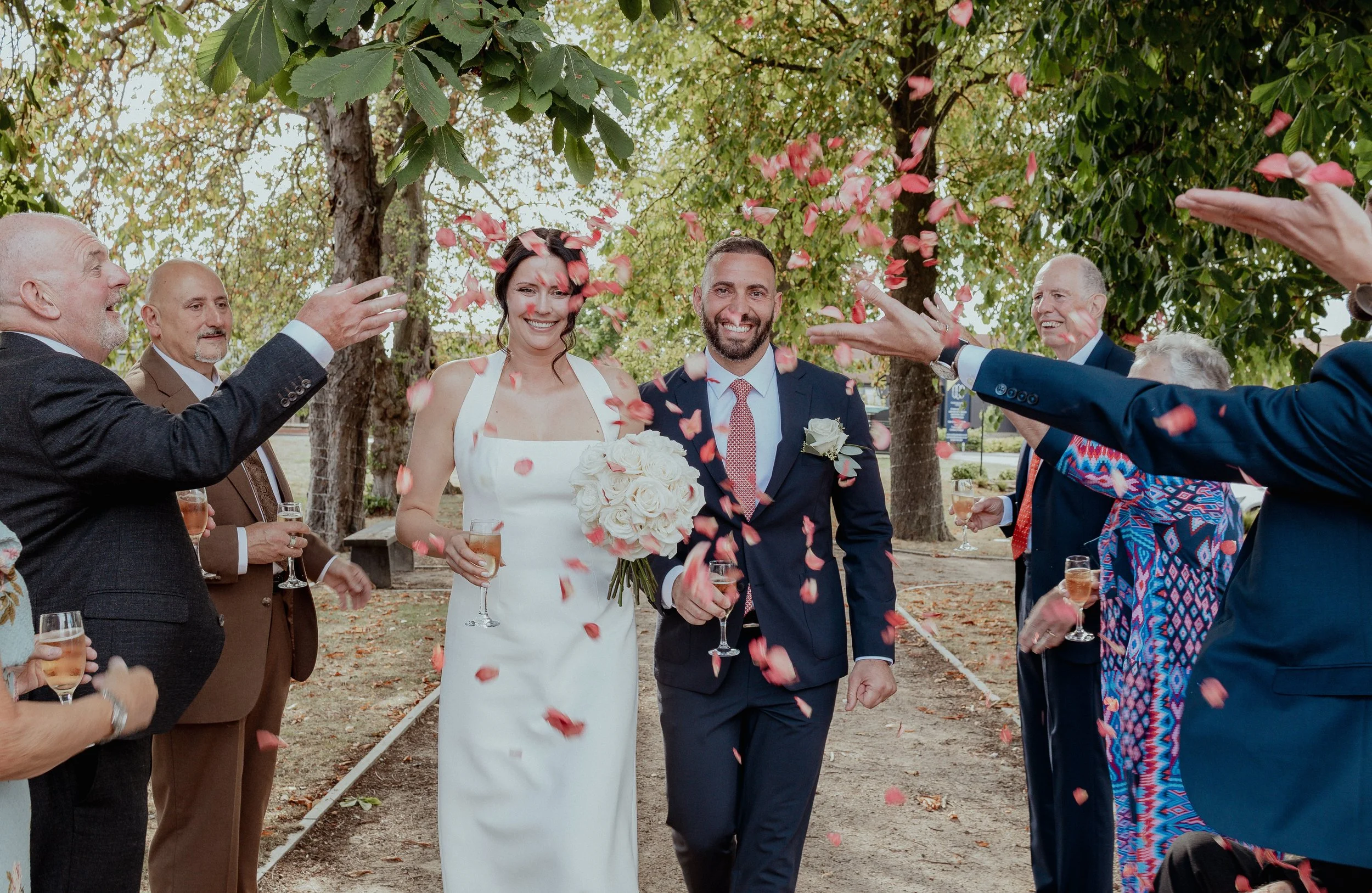Bride and groom walking through confetti during wedding celebration outside, surrounded by friends and family.