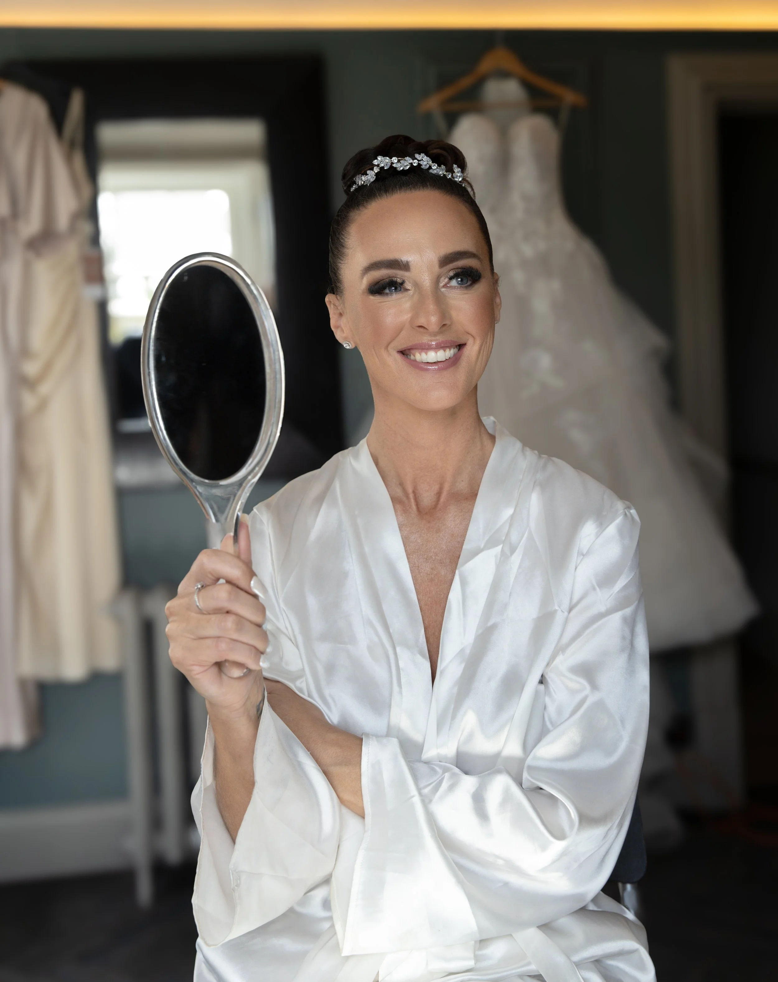 Brunette woman in satin robe with pearl headband smiling at mirror in bridal suite with wedding dress hanging in the background.