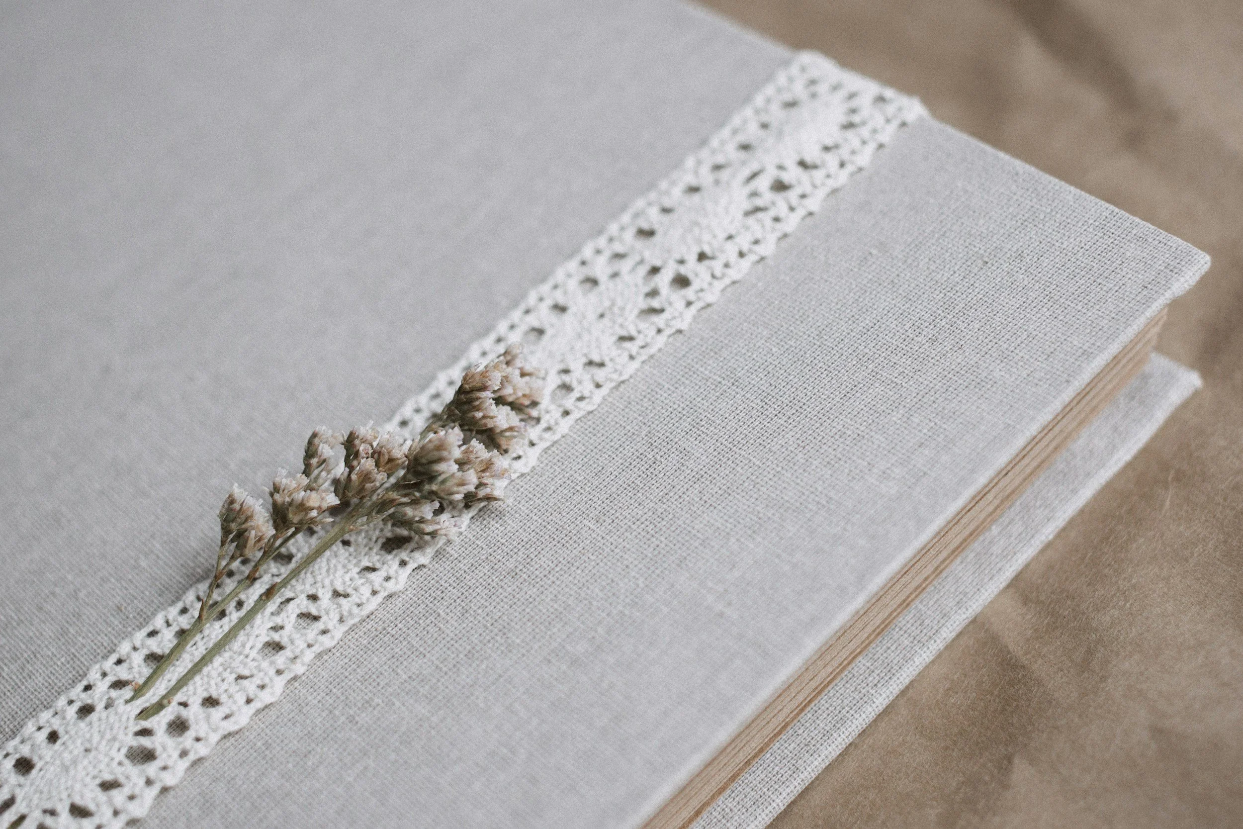 A gray book with a lace ribbon and dried flowers on top, resting on a tan surface.