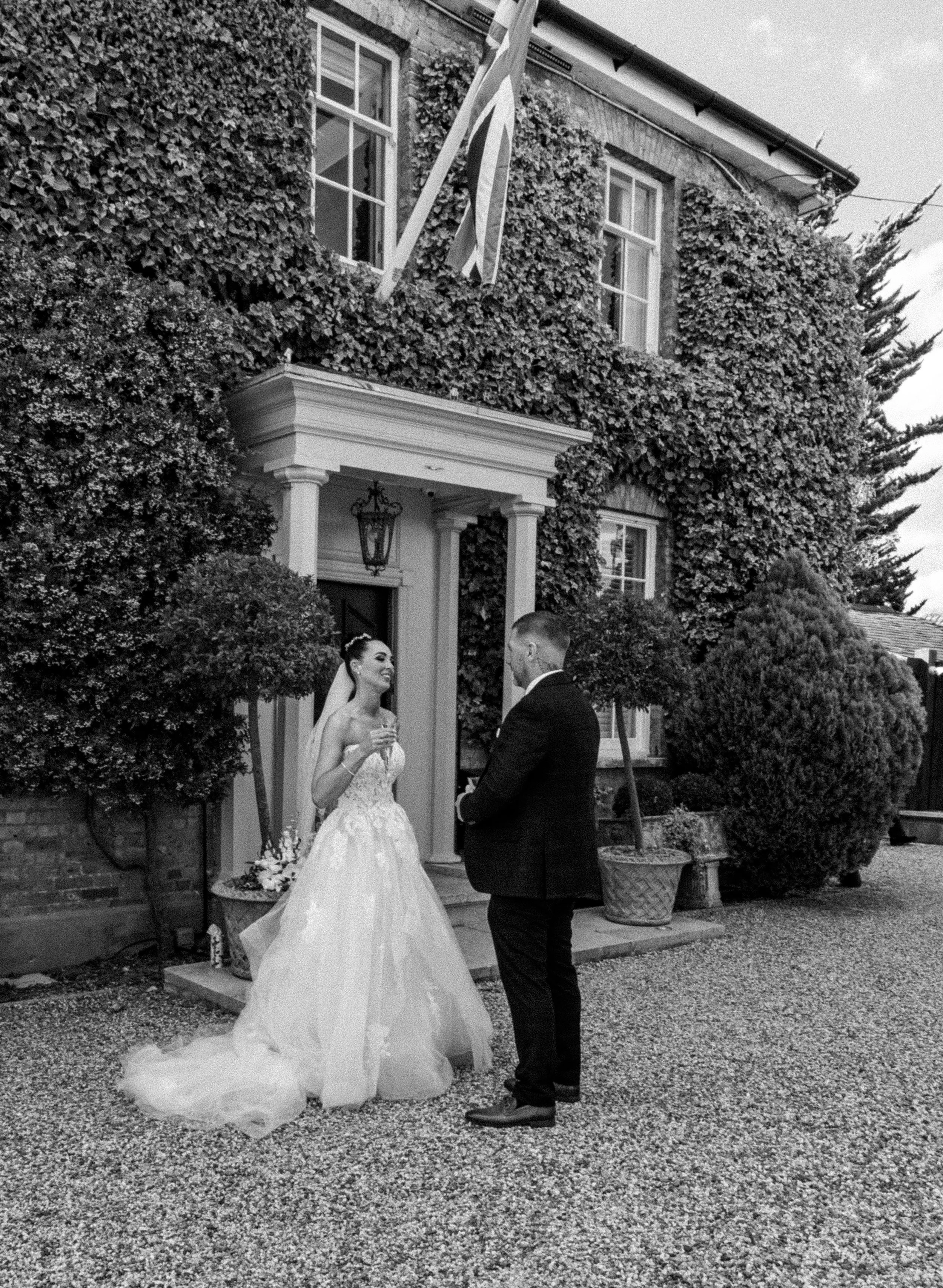 A bride and groom standing outside a house, engaged in a conversation. The bride is in a wedding dress, holding a glass, and smiling. The house has ivy-covered walls, a front porch with columns, and a Union Jack flag hanging. There are trees and shru
