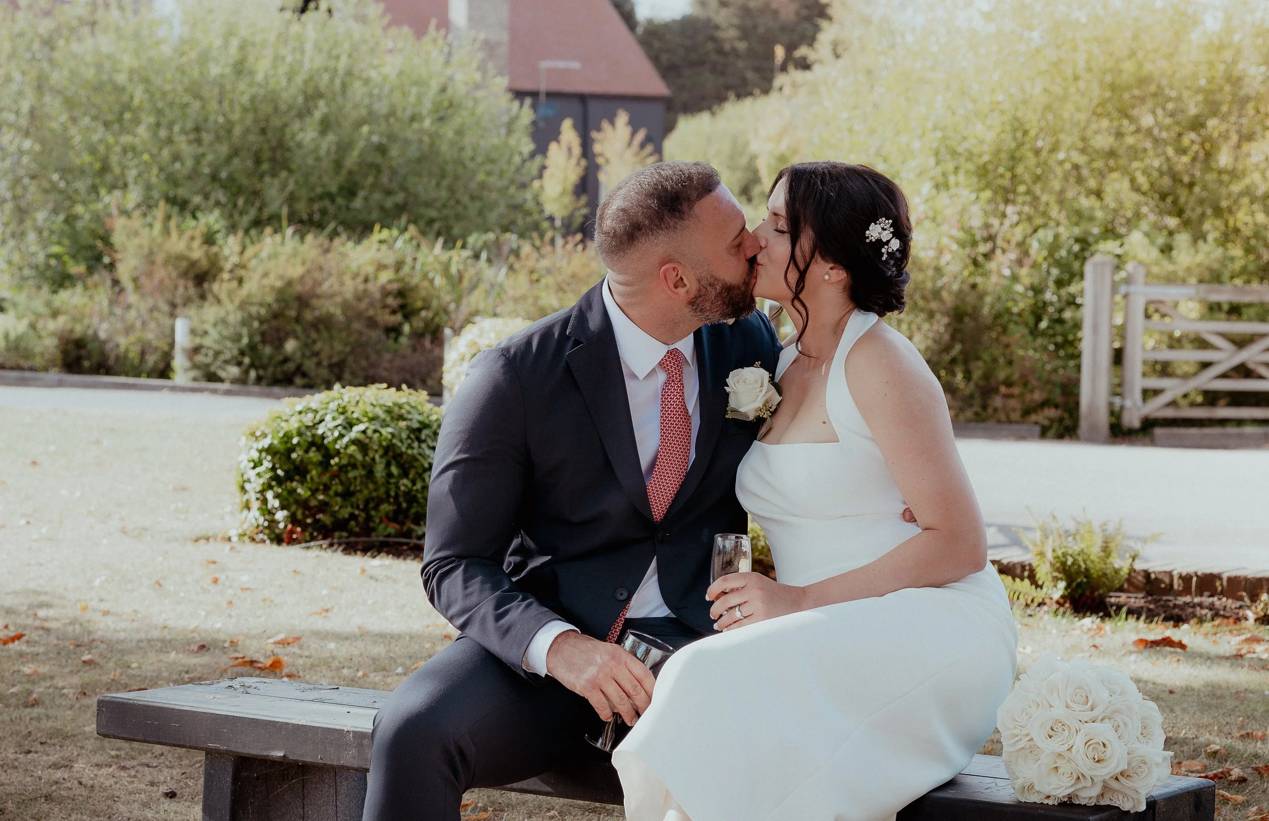A newlywed couple shares a kiss while sitting on a bench in a garden surrounded by greenery, with the bride holding a glass of champagne and a bouquet of white roses resting on the bench nearby.