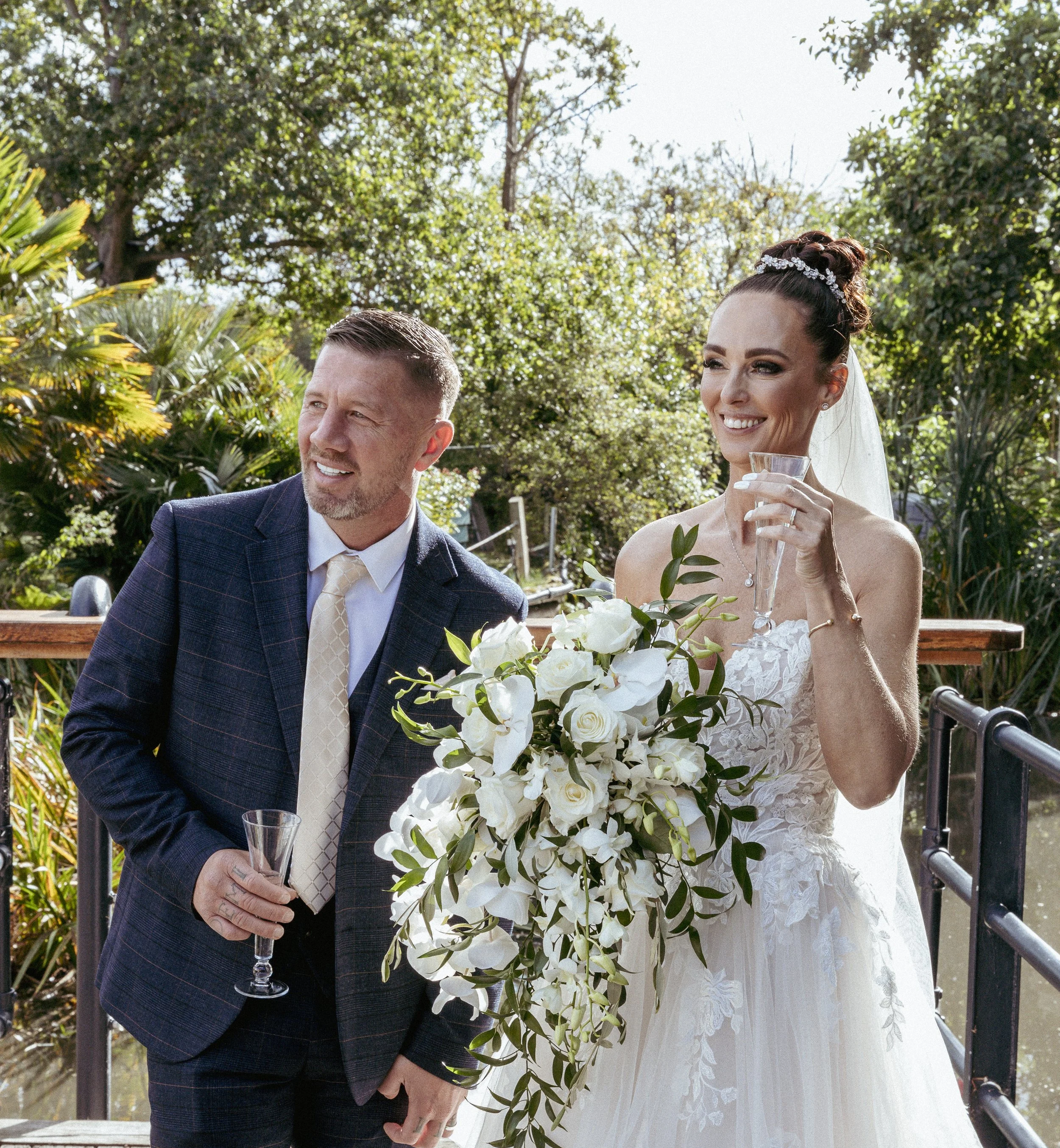 A bride and a man, possibly her father, are outdoors at a wedding celebration, holding champagne glasses, with lush greenery in the background.