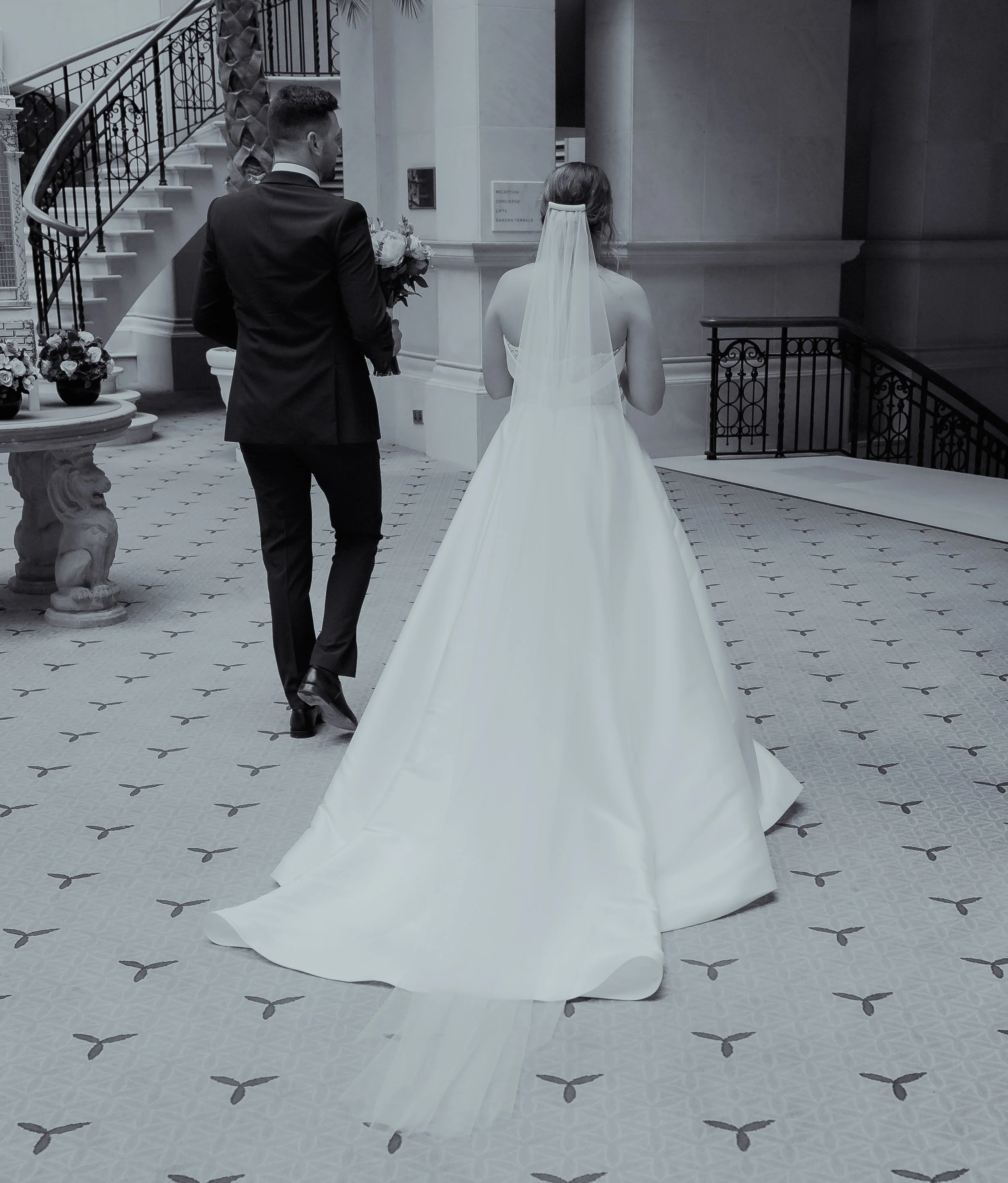 Bride and groom walking indoors, bride in a long white wedding gown with veil, groom in a black suit, holding flowers, staircase and ornate details visible.