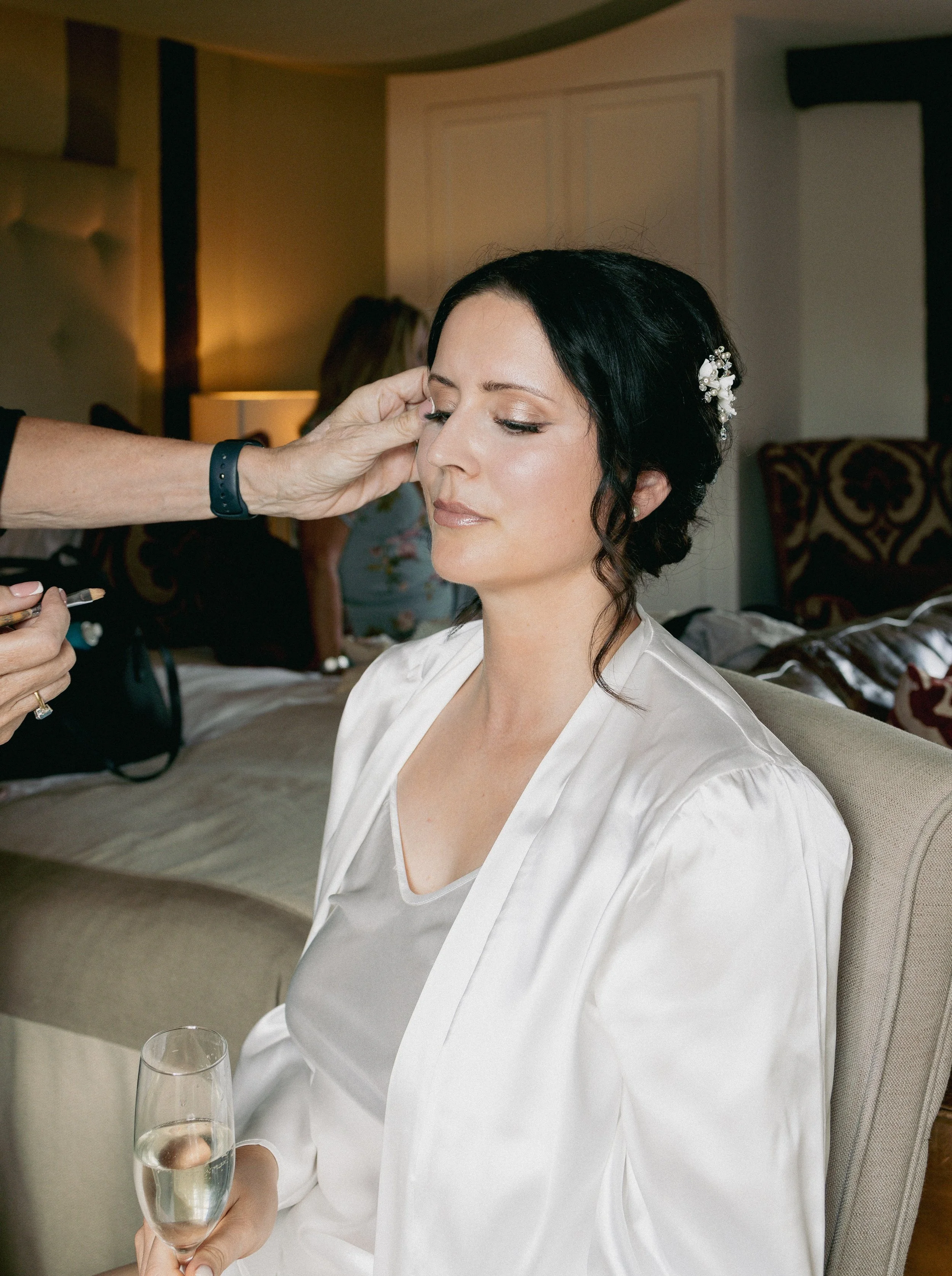 A woman with dark hair and light makeup sitting with a glass of champagne, having her makeup touched up by a makeup artist, in a room with warm lighting and patterned pillows.