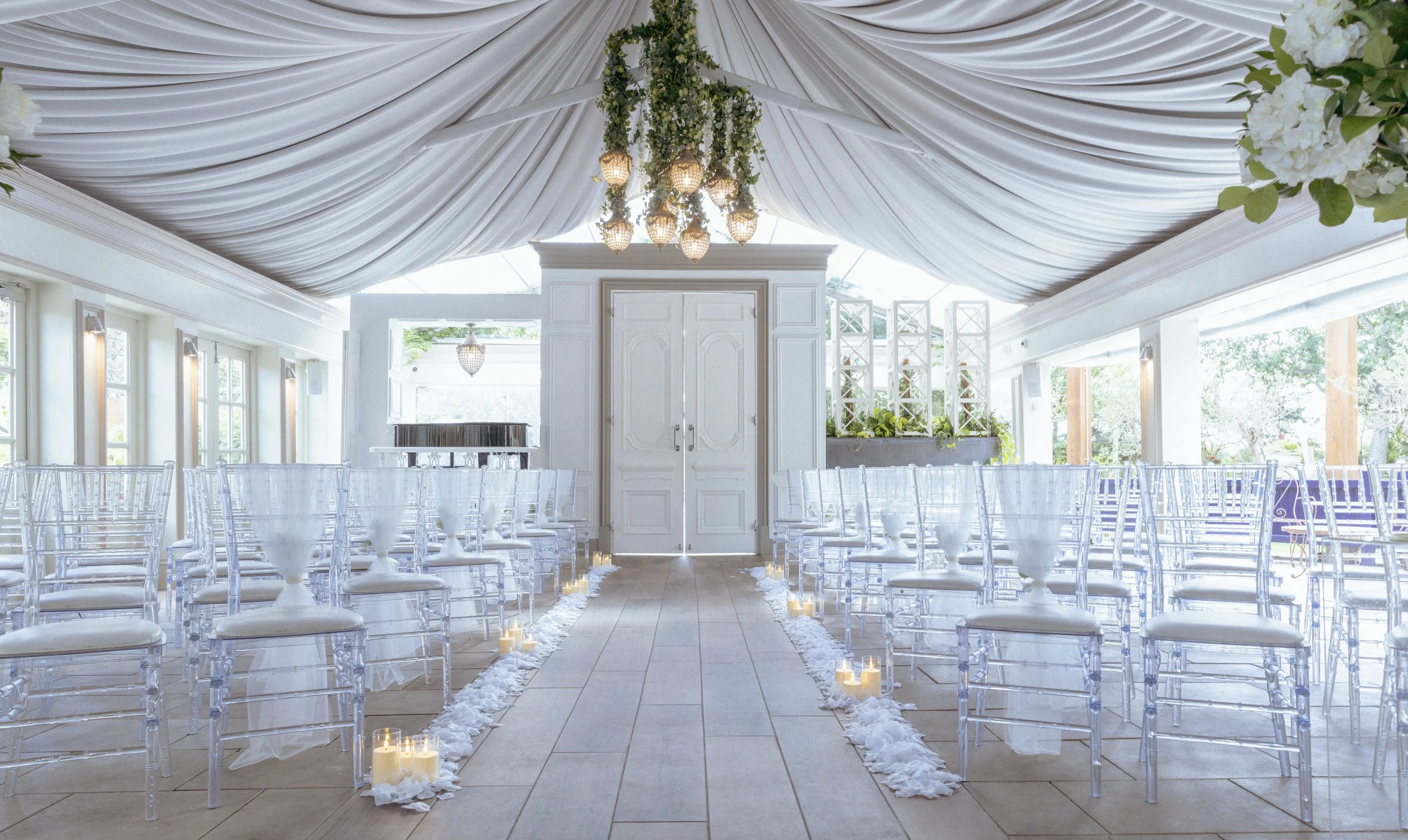 Elegant white wedding ceremony setup inside a tent with transparent chairs, aisle decorated with candles and white fabric, and a canopy of white drapery overhead.
