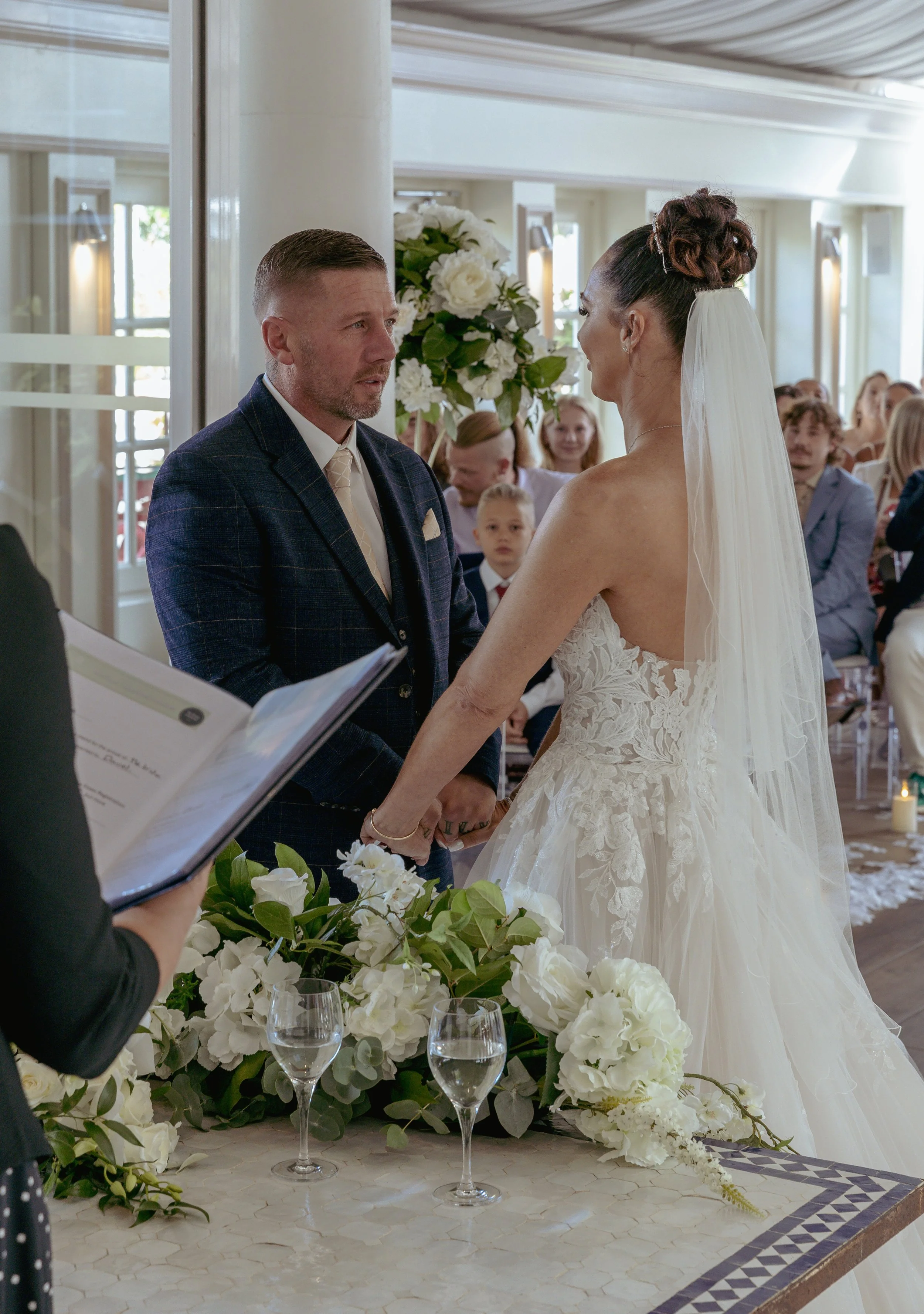 A bride and groom holding hands during their wedding ceremony, with guests seated in the background and a floral arrangement on the table in front of them.