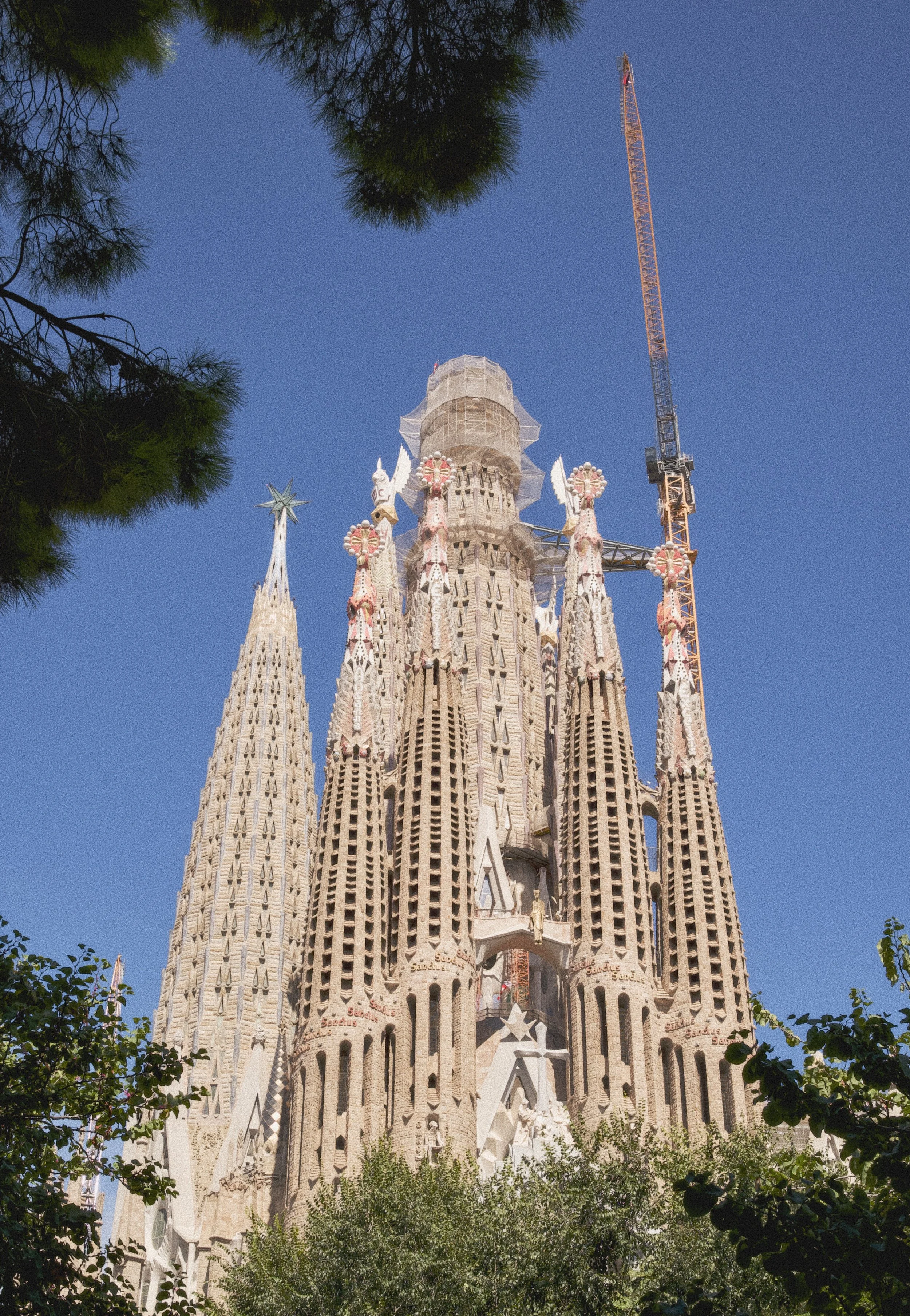Sagrada Familia basilica under construction in Barcelona, Spain, with a crane, trees in the foreground, and a clear blue sky.