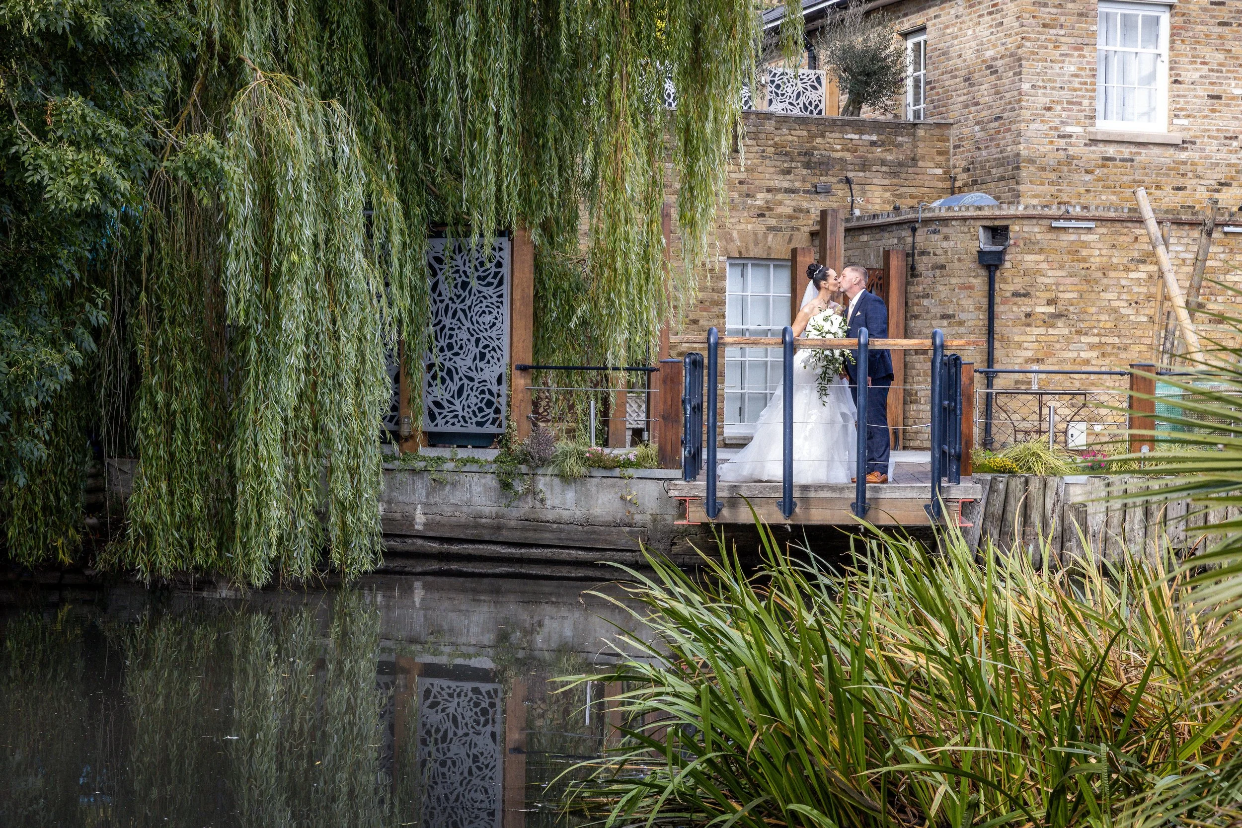A bride and groom sharing a kiss on a small wooden bridge over a canal, with lush green trees and plants around them, and old brick buildings in the background.