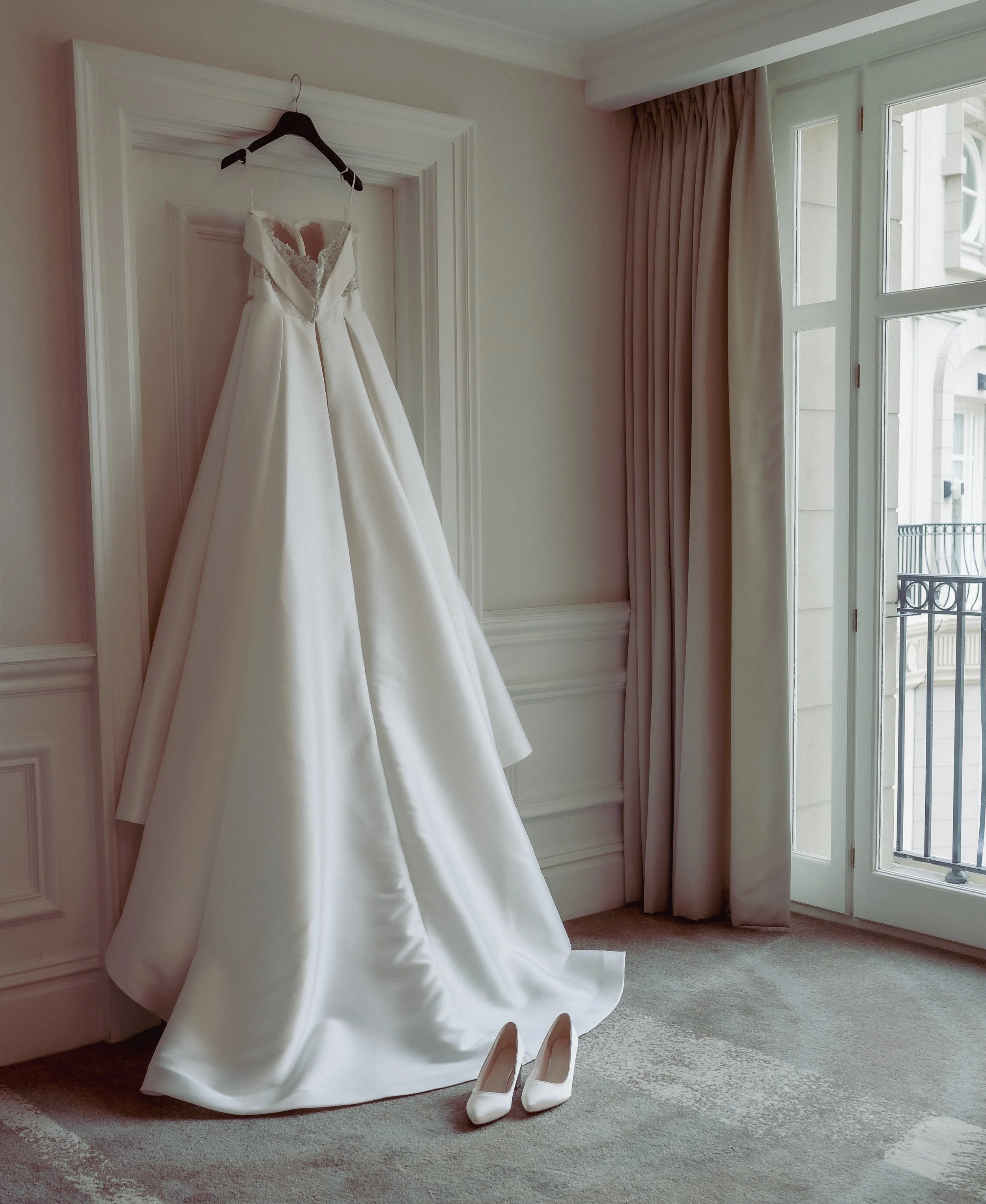 A white wedding dress hanging on a black hanger, with white high heels placed on the floor underneath, in a room with light-colored walls, curtains, and a window showing a balcony outside.