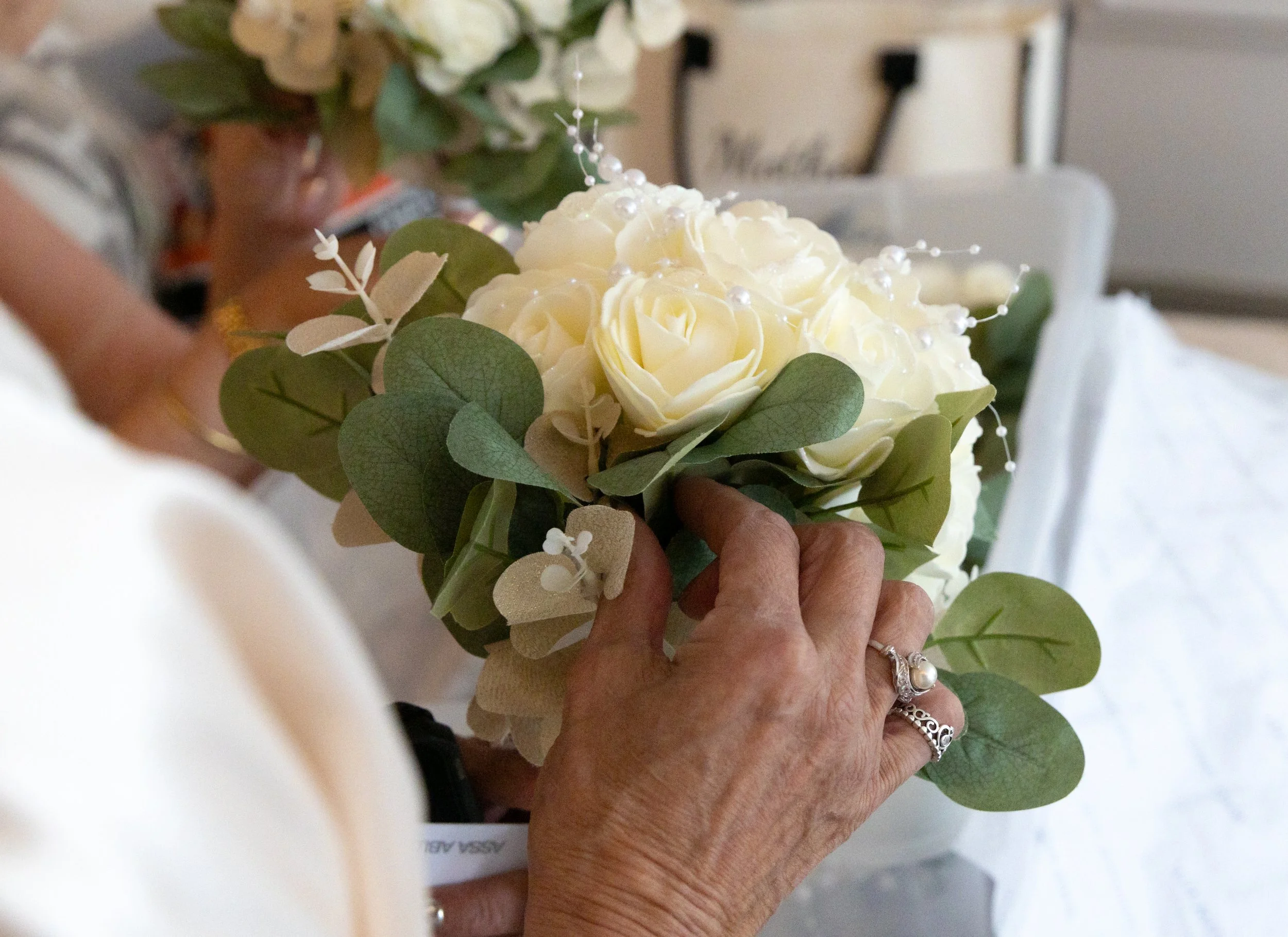 Close-up of a person holding a bouquet of white roses and green leaves, with vintage rings on their finger.