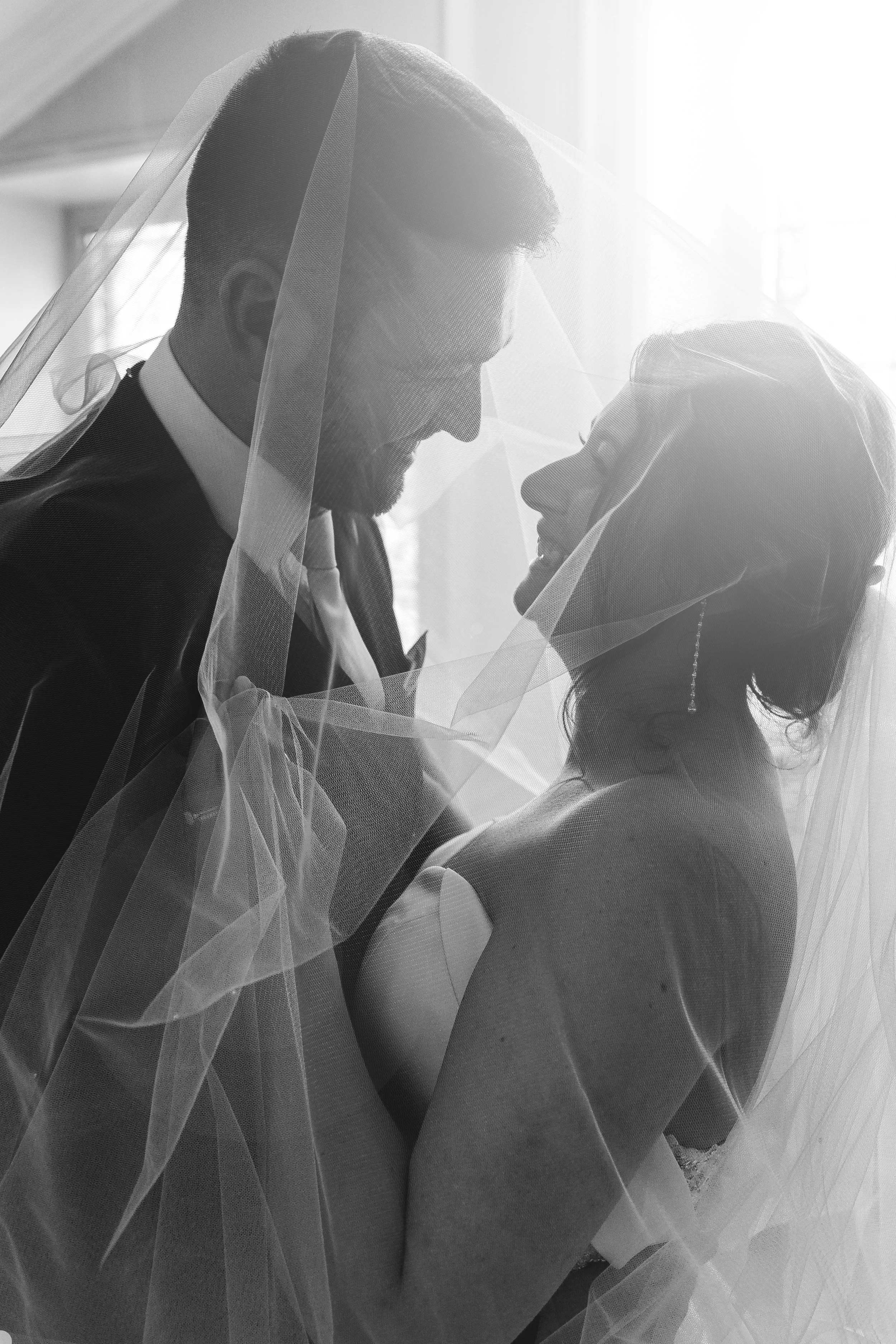 Black and white photo of a bride and groom smiling at each other, standing close with a veil draped over both of them, in a bright room.
