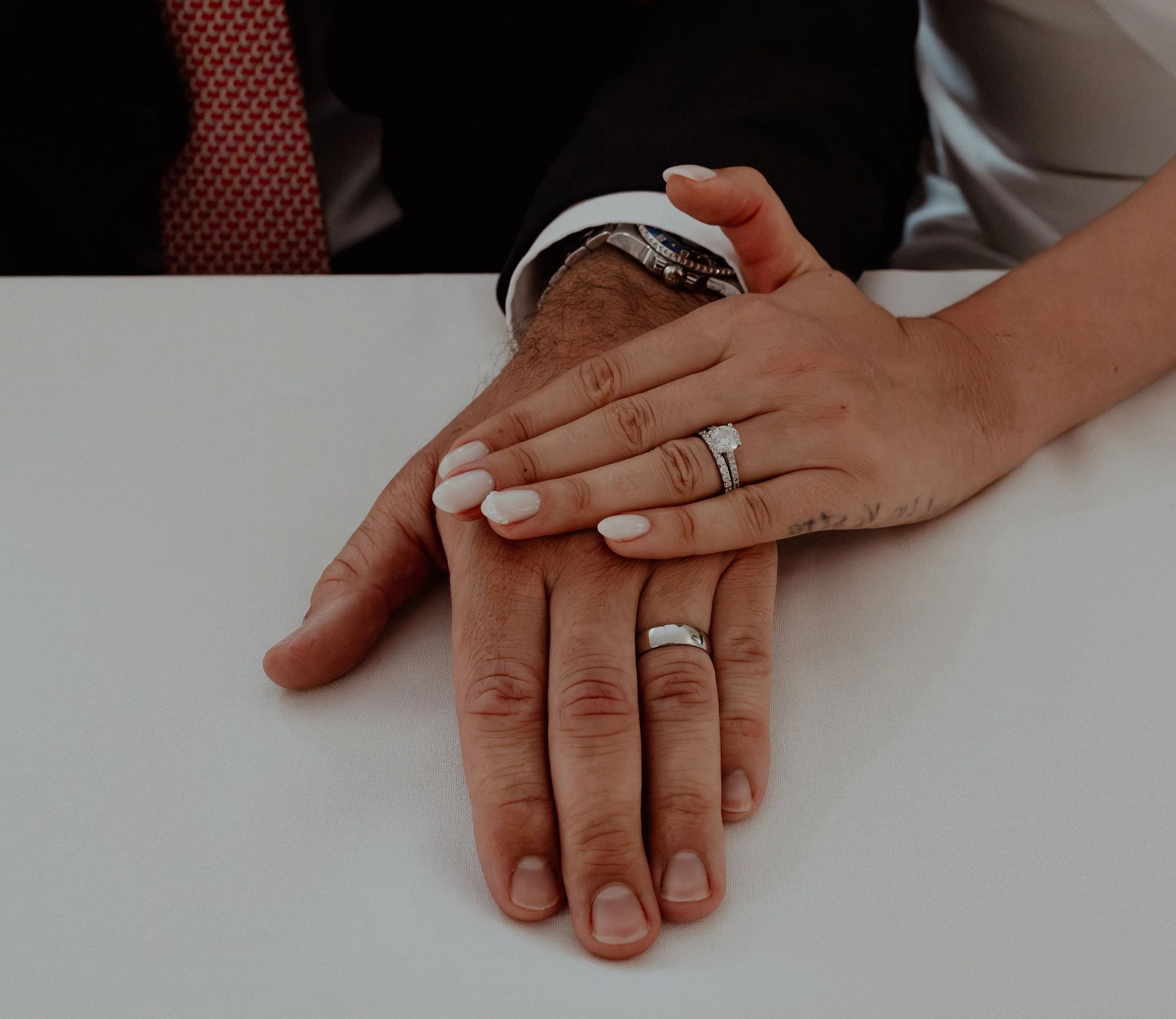 Close-up of a man and woman holding hands on a white table, both showing wedding rings.