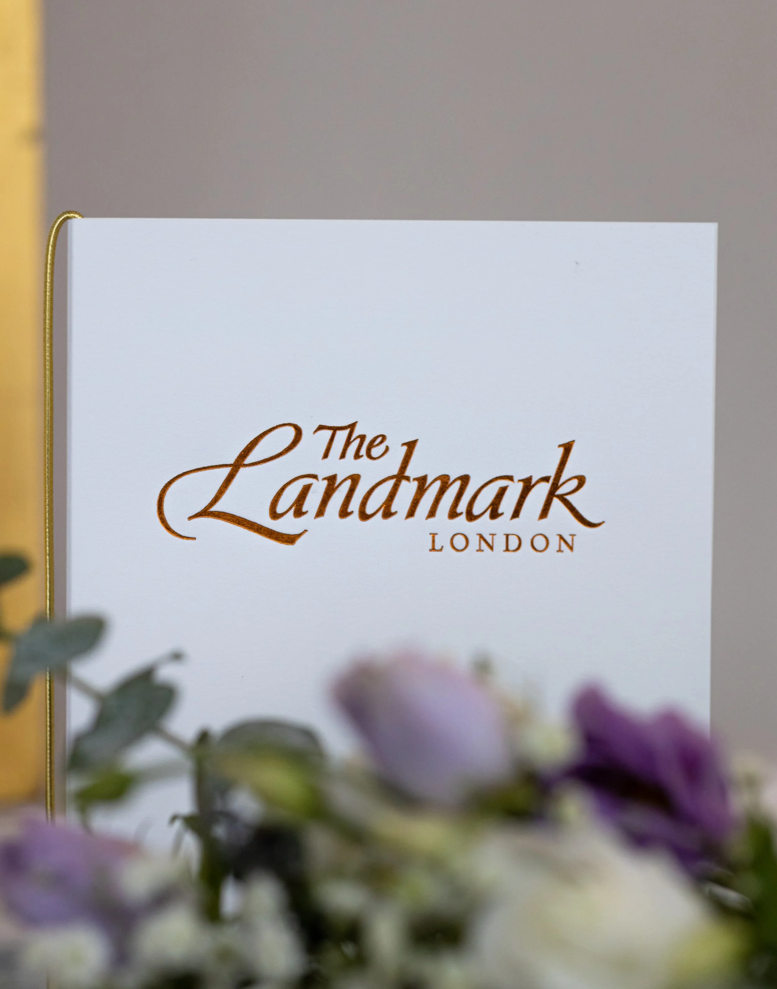 Close-up of a white card with gold cursive text reading 'The Landmark London' and a blurred floral arrangement in the foreground.