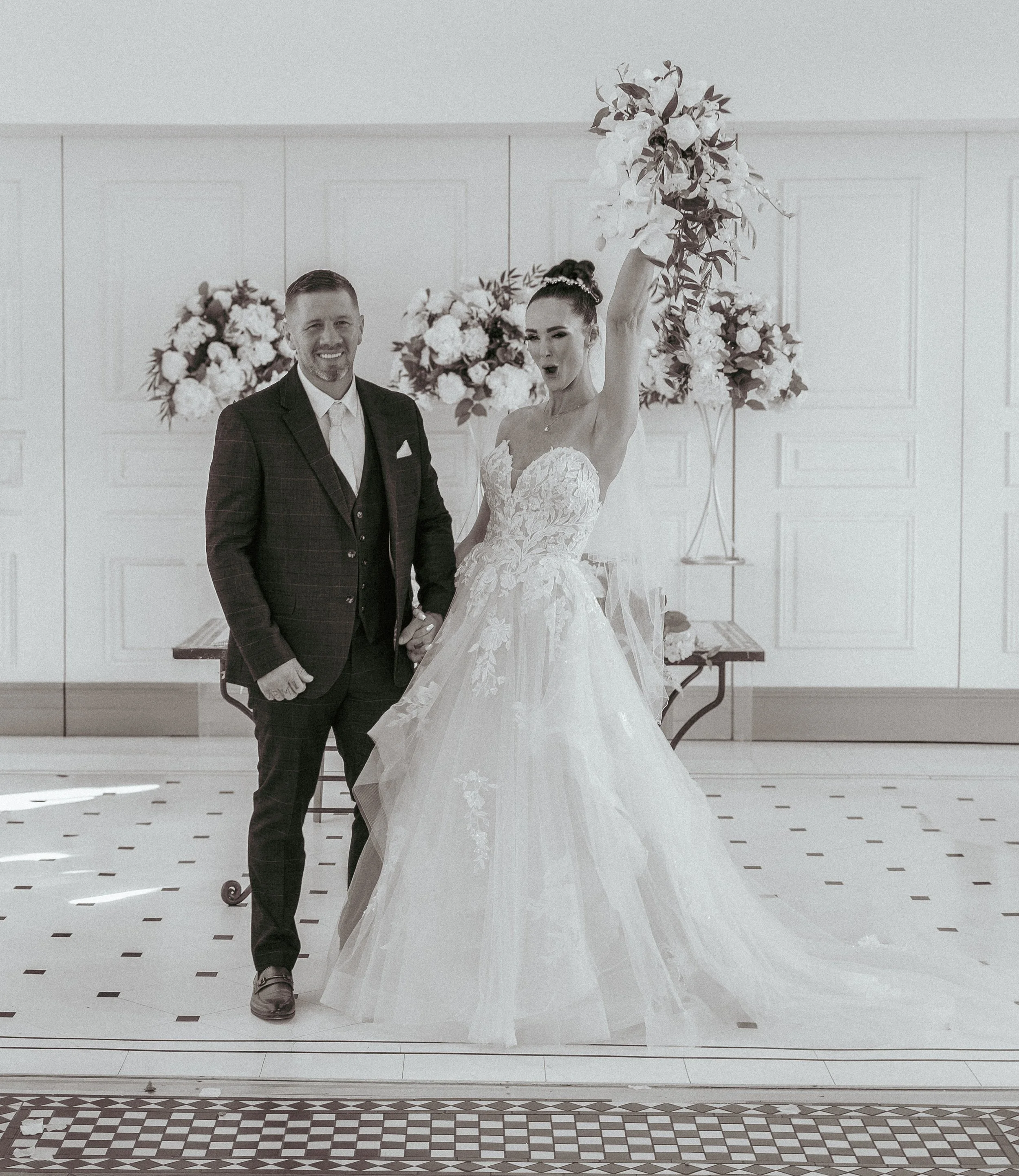 A bride and groom standing together at their wedding. The bride is holding a bouquet of flowers raised in the air, and the groom is smiling while holding her hand. They are inside a decorated hall with floral arrangements.
