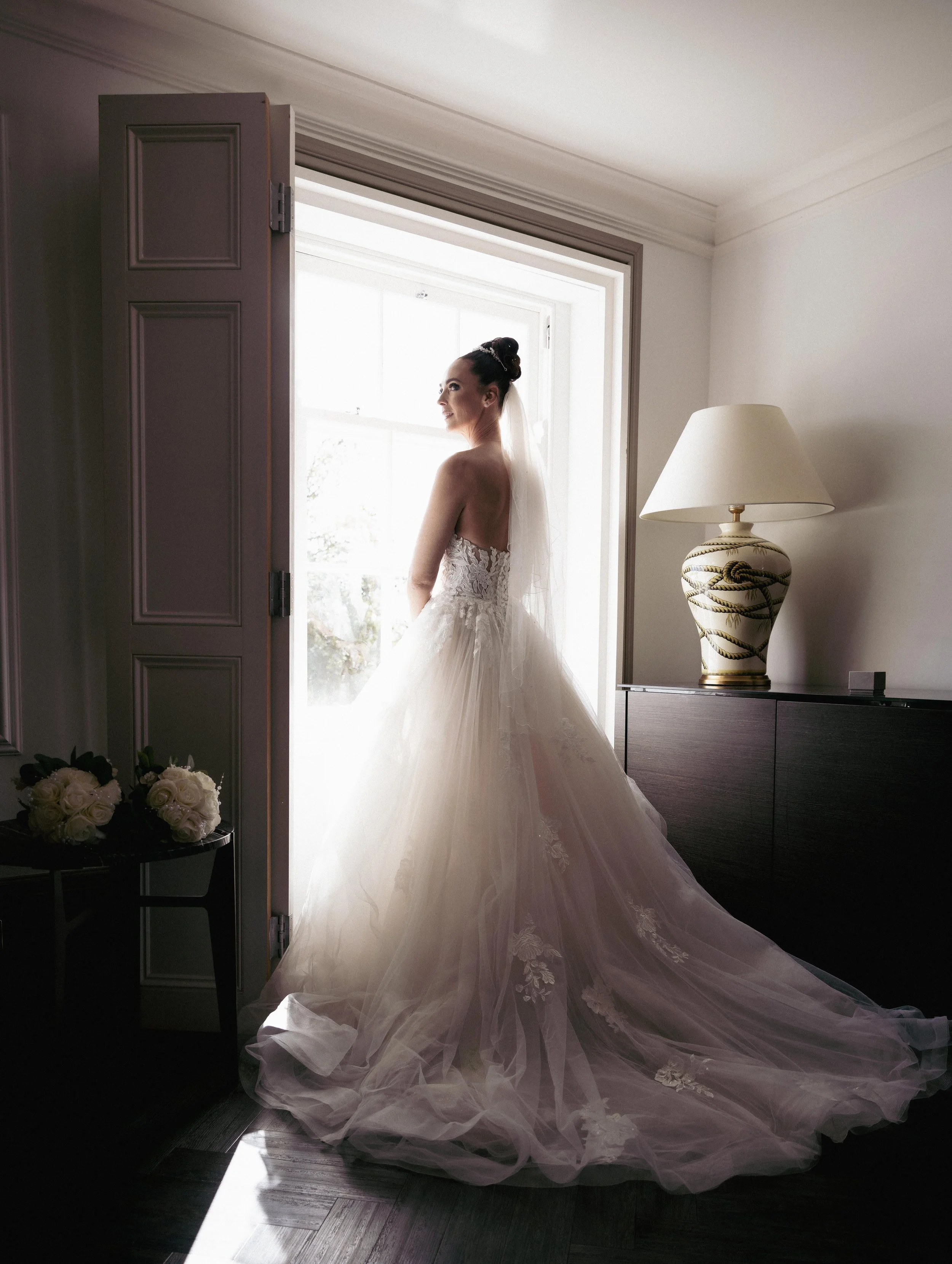 A bride in a wedding dress standing by a window, looking outside, with sunlight illuminating her.