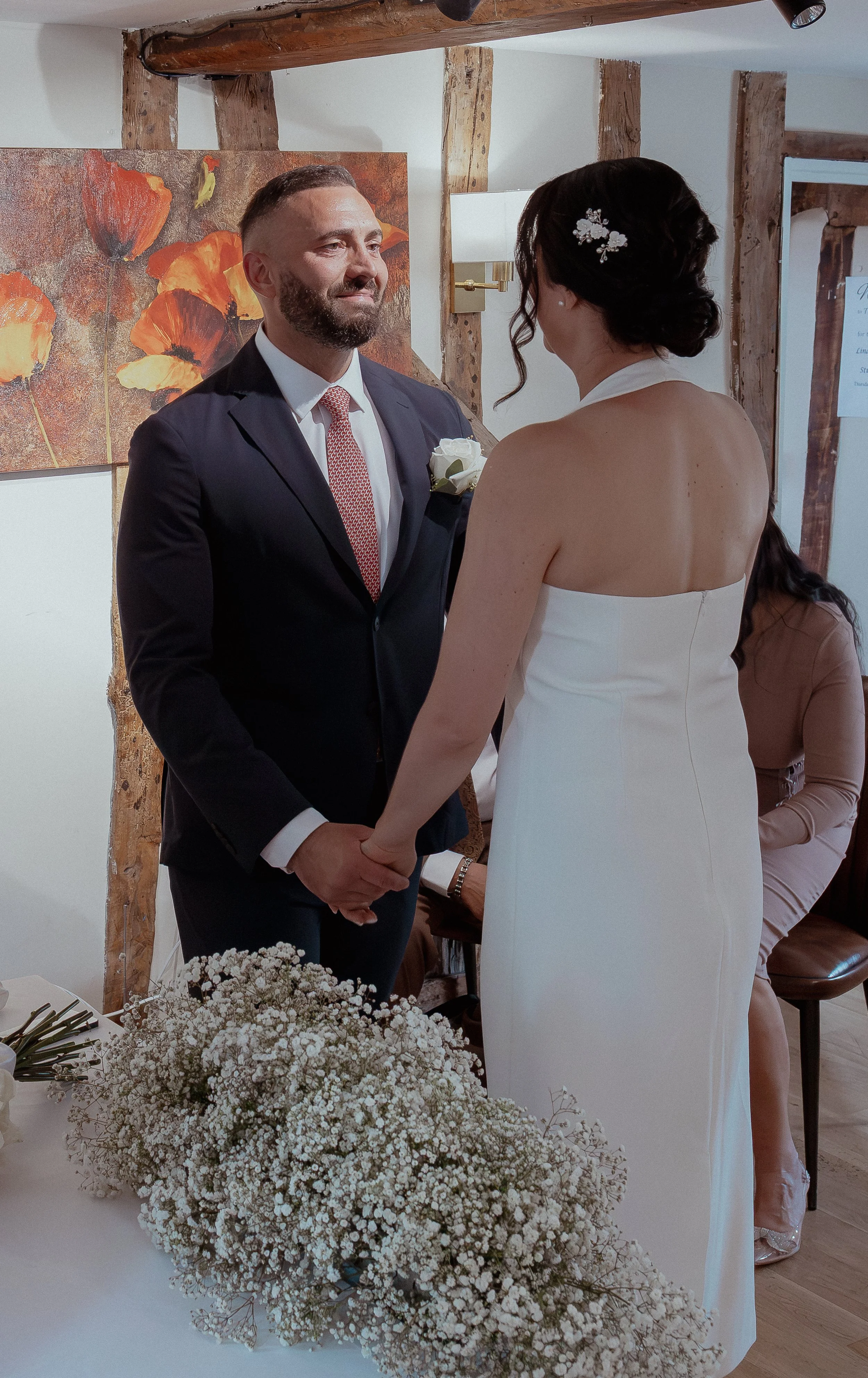 A bride and groom holding hands during their wedding ceremony indoors, with flowers on the table in front of them and a painting of orange flowers on the wall behind.