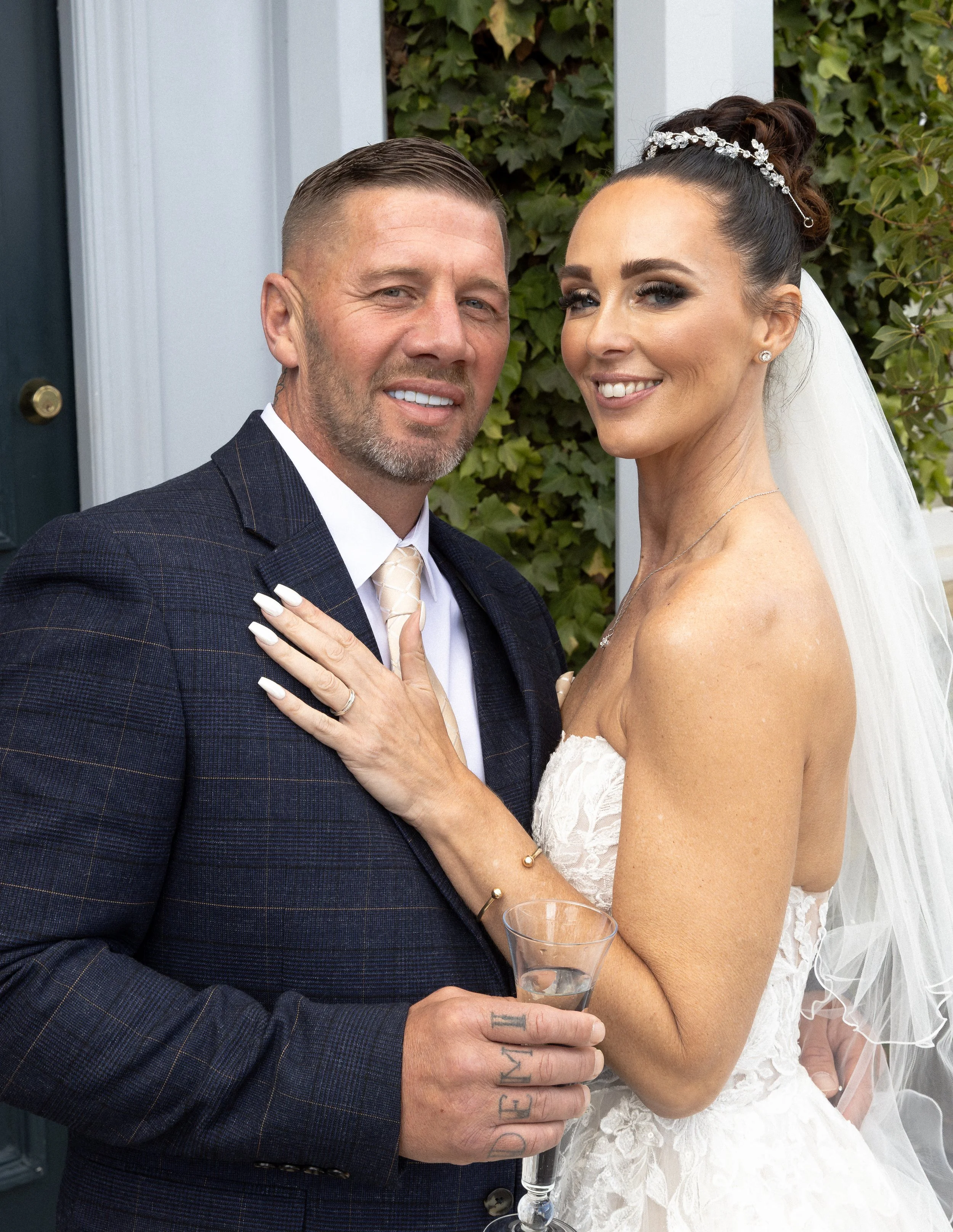 A couple in wedding attire smiling and posing for a photo, with the bride wearing a white dress and veil, and the groom in a suit holding a glass of champagne.