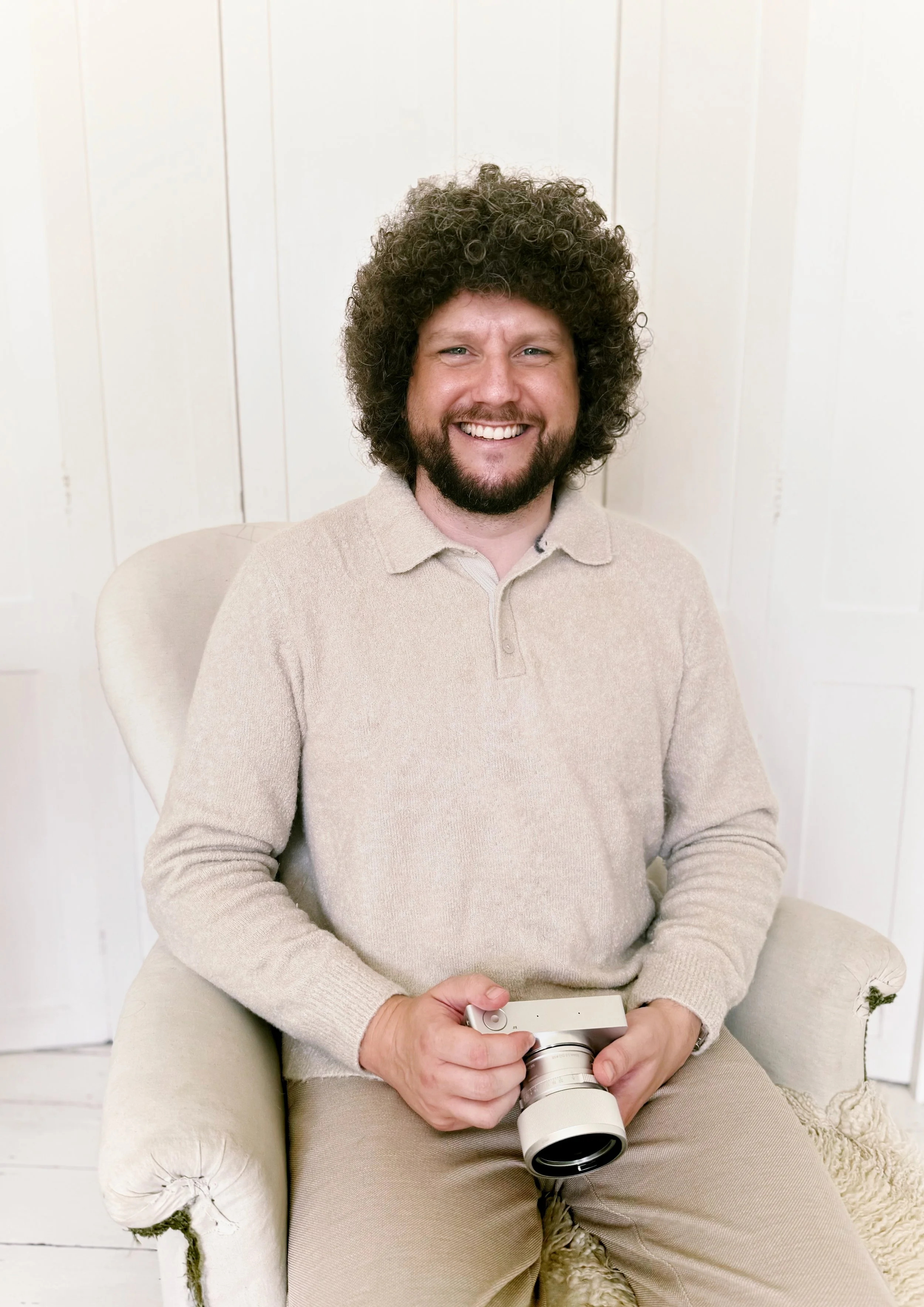 Smiling man with curly hair and beard holding a camera, sitting on a light-colored armchair in a room with white walls.
