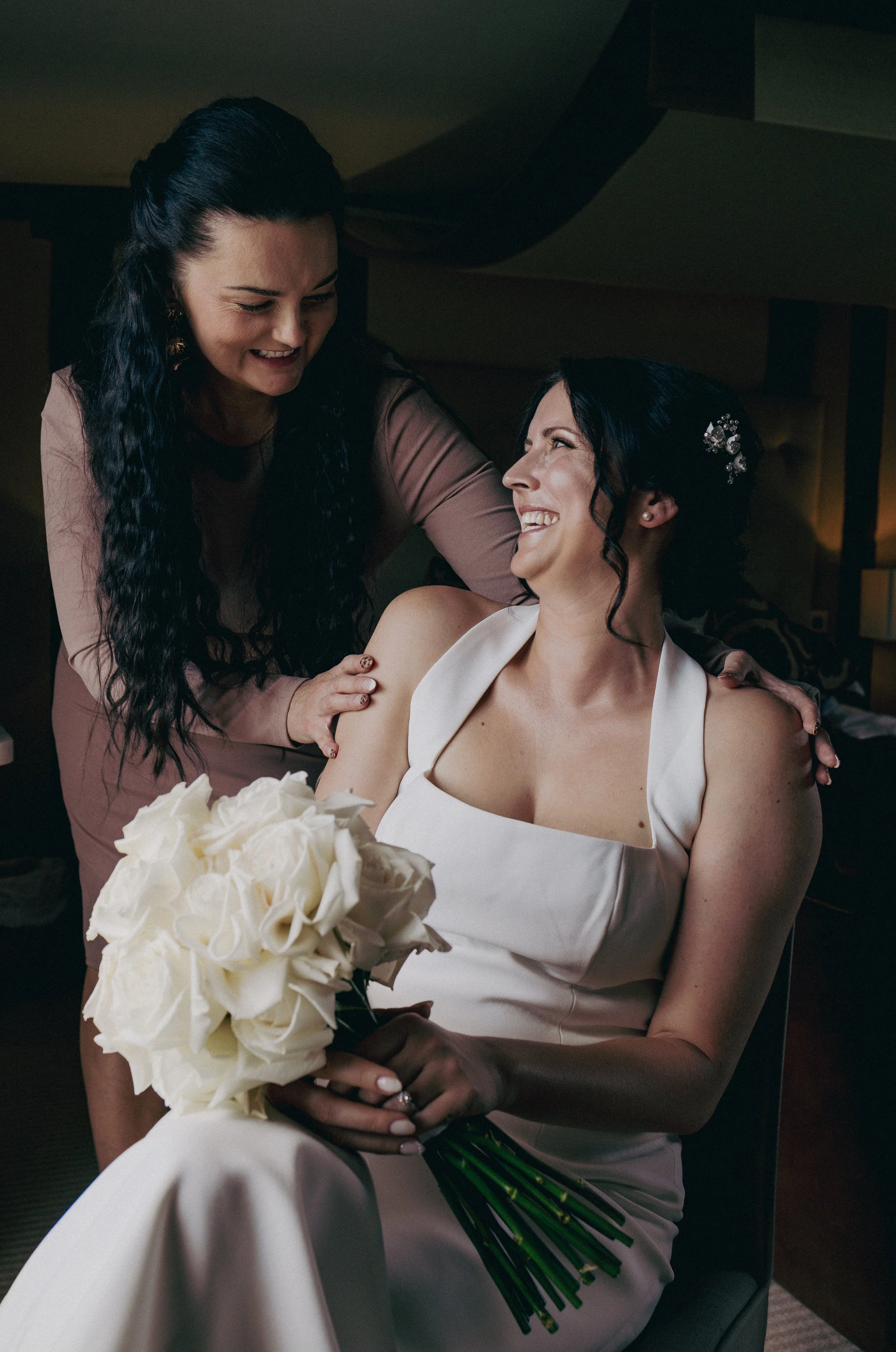 A bride in a white dress holding a bouquet of white roses, smiling at a woman helping her prepare, in a warmly-lit room.