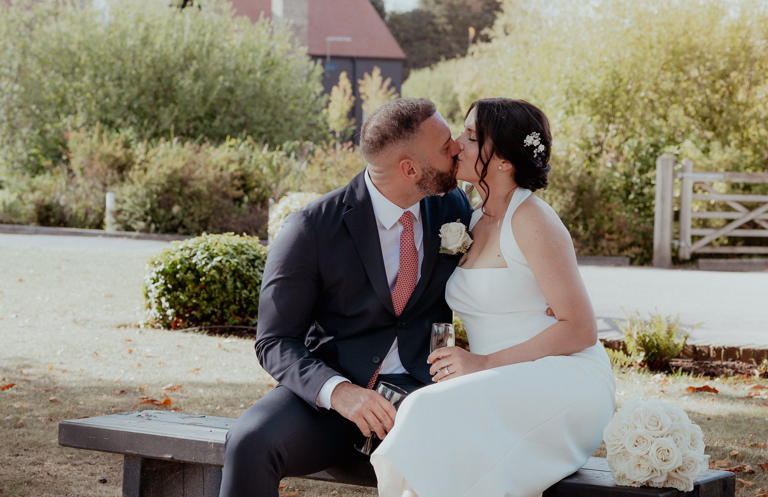 A newlywed couple sharing a kiss outdoors, seated on a wooden bench with a bouquet of white roses beside them. The bride is wearing a white wedding dress with floral hair accessories, and the groom in a black suit with a white shirt and red tie, holding a glass of champagne.