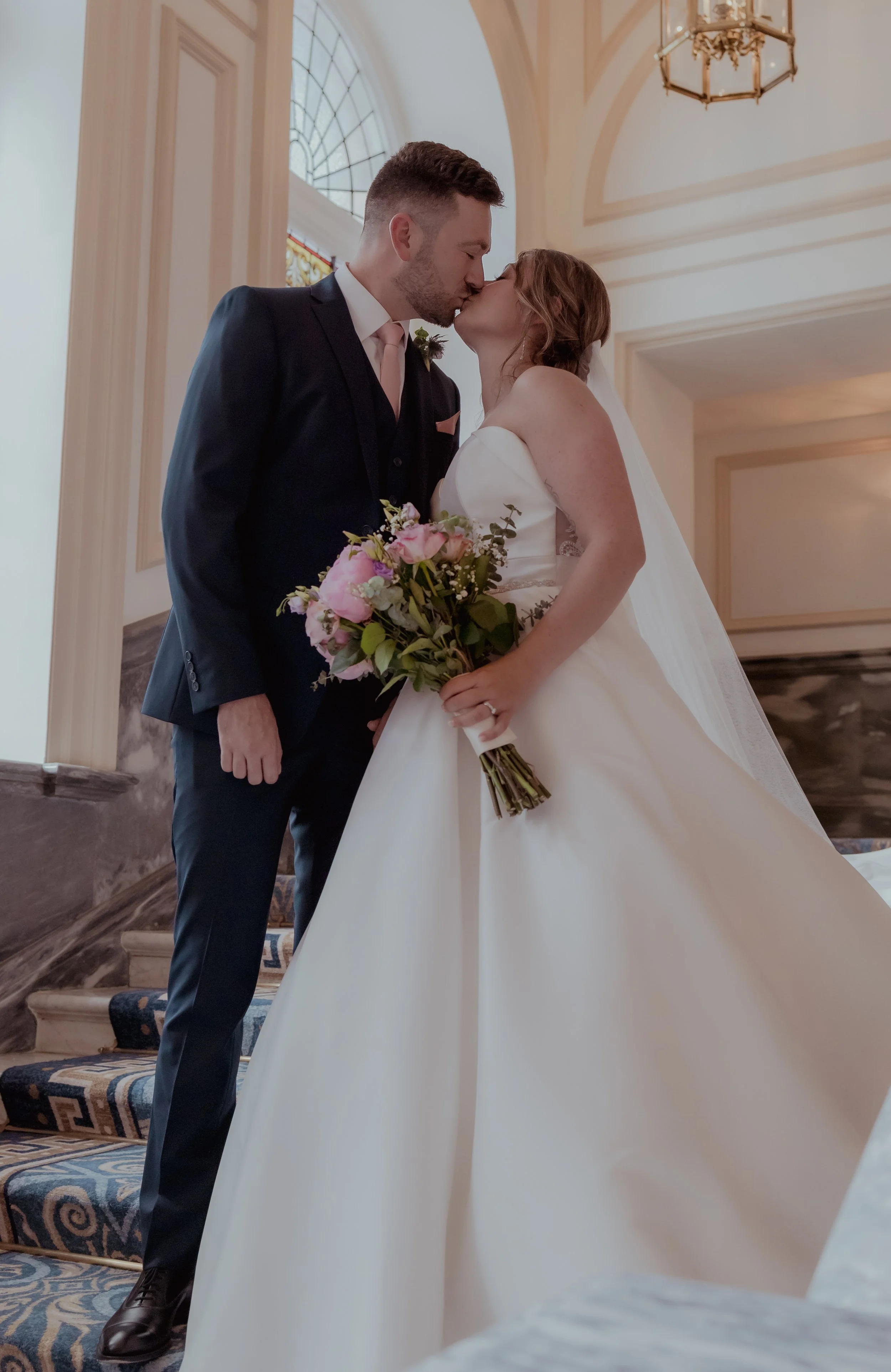A bride and groom sharing a kiss inside a church or wedding venue, with the bride holding a bouquet of pink and white flowers, and the groom wearing a black suit and pink tie.