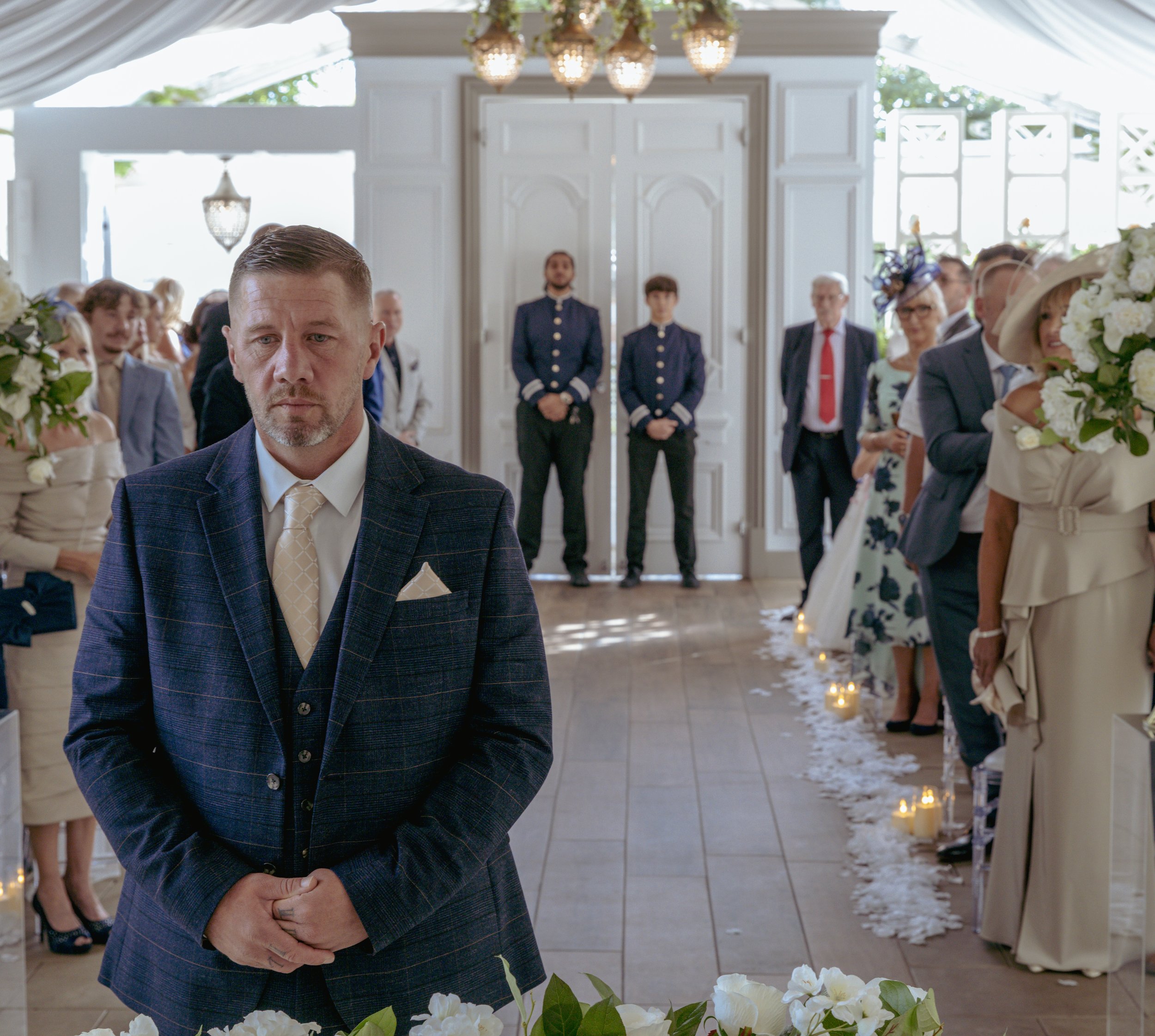 A man stands alone at the front of a wedding ceremony, wearing a dark plaid suit, with a serious expression. Guests are standing on either side of a floral aisle, some holding bouquets and wearing formal attire, in a bright, decorated indoor venue wi