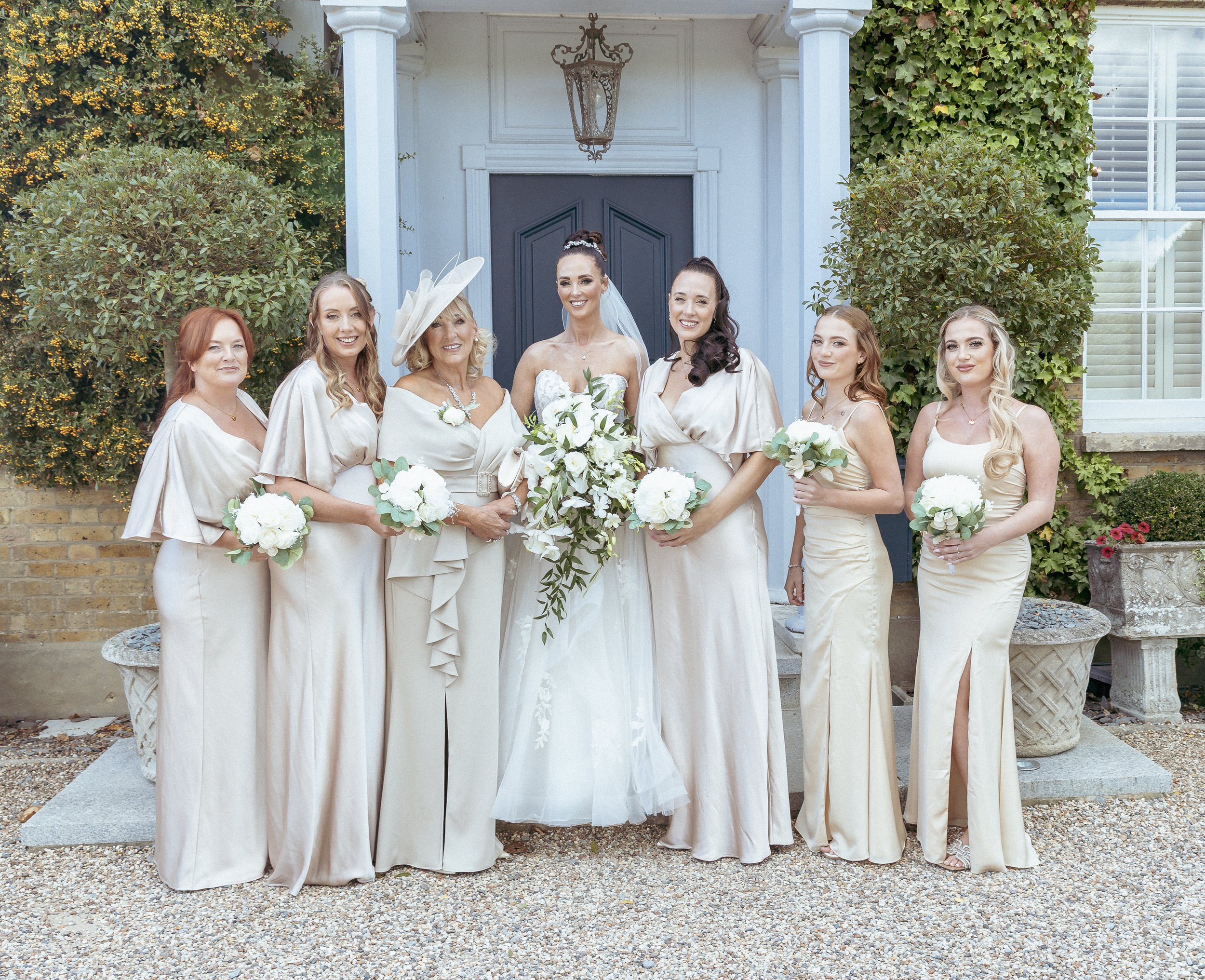 A bride and seven women, likely bridesmaids and a mother of the bride, standing in front of a house with ivy-covered walls and a blue door, all dressed in cream-colored dresses and holding white bouquets.