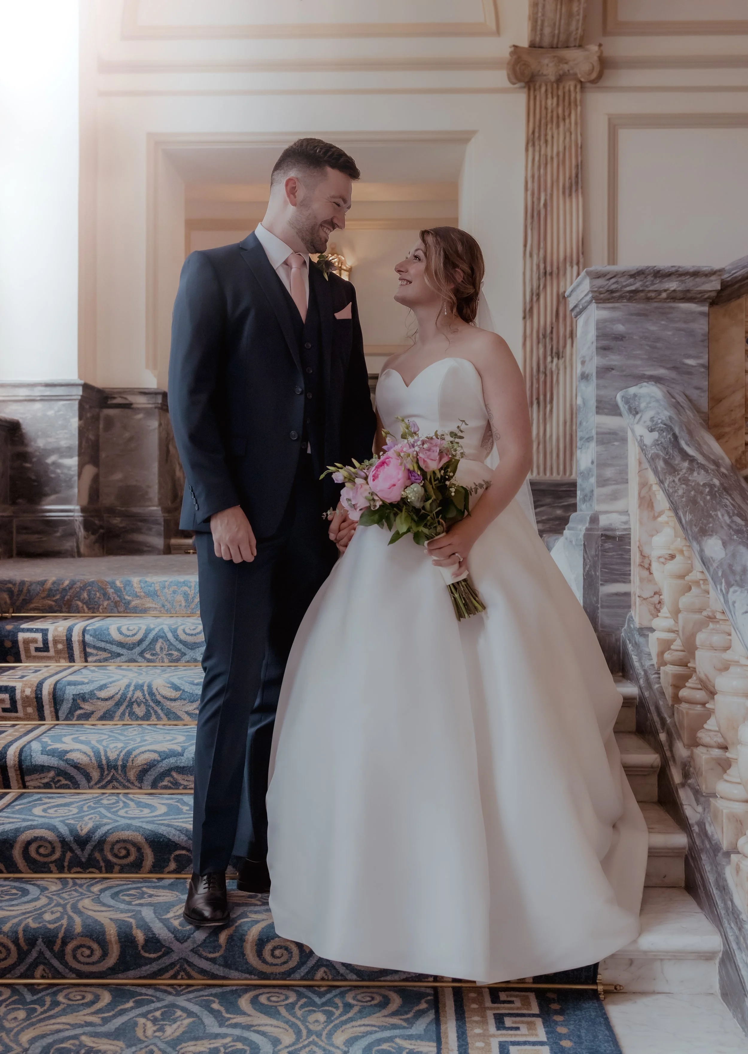 A bride and groom standing on stairs indoors, smiling and gazing at each other. The bride wears a strapless white wedding gown and holds a bouquet of pink and white flowers. The groom wears a dark suit with a light pink tie and boutonniere.