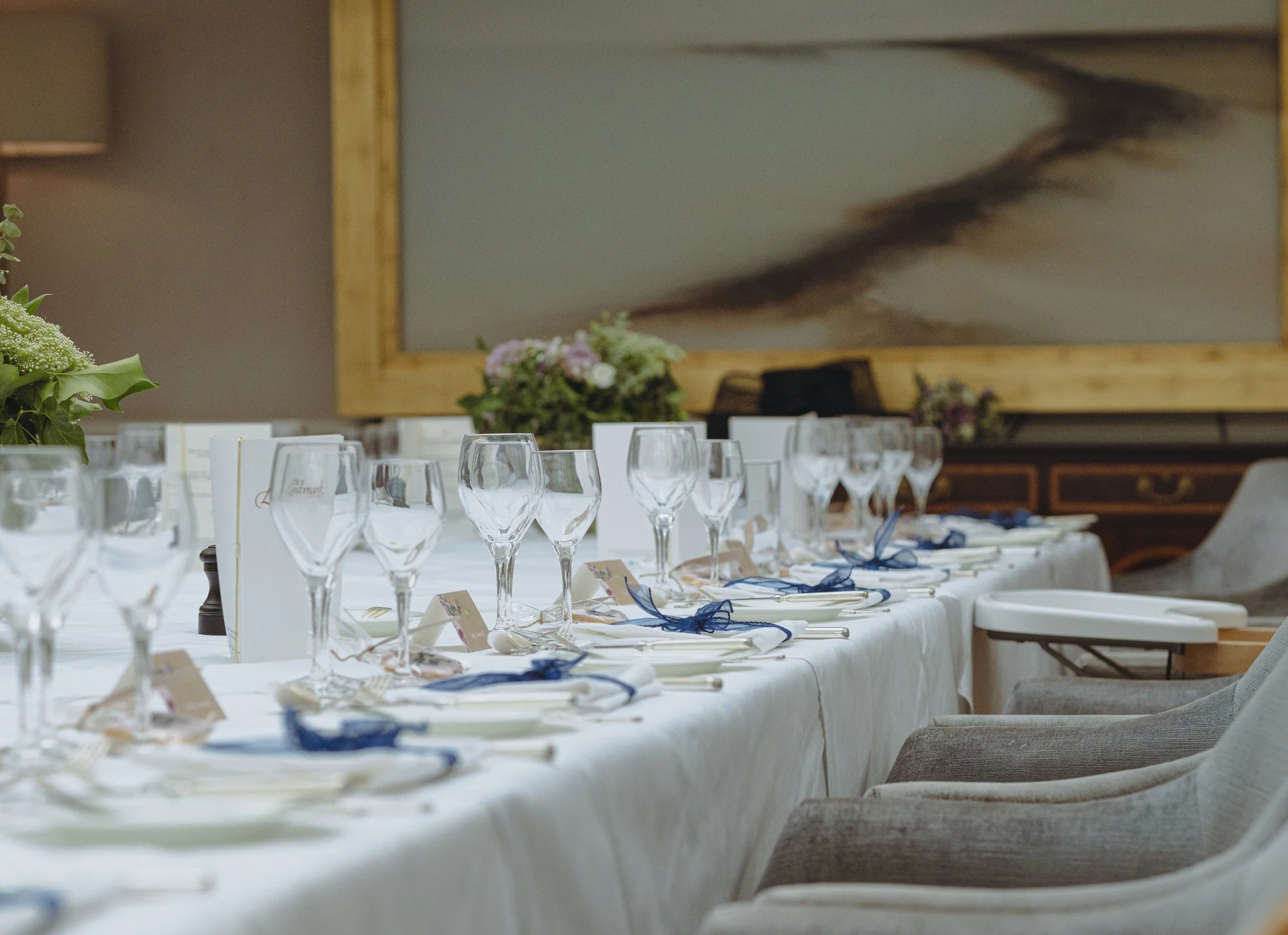 Elegant dining table set with wine glasses, silverware adorned with blue ribbons, and floral arrangements in the background.