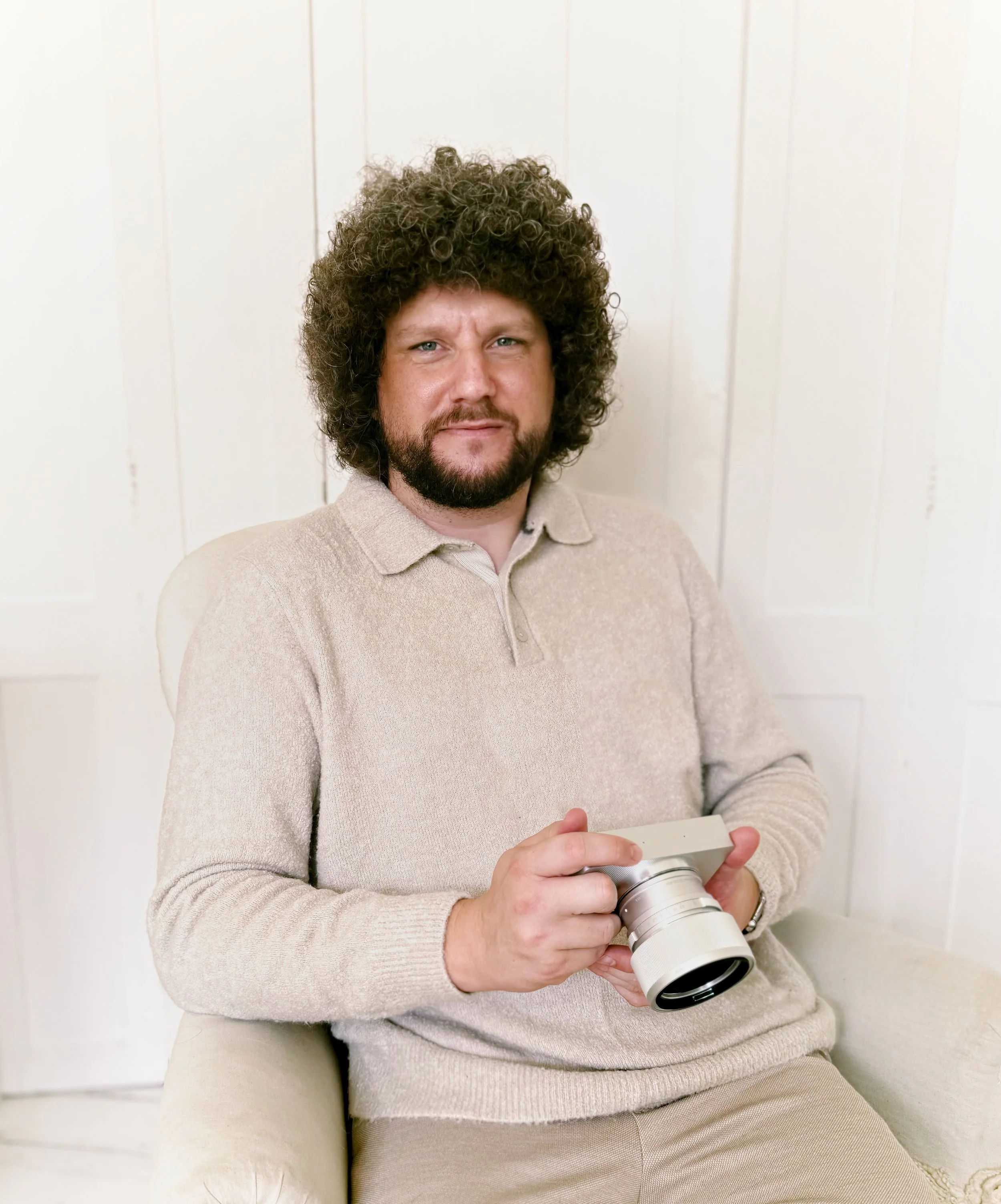 Man with curly hair and beard holding a camera, sitting on a beige chair against white wooden wall.