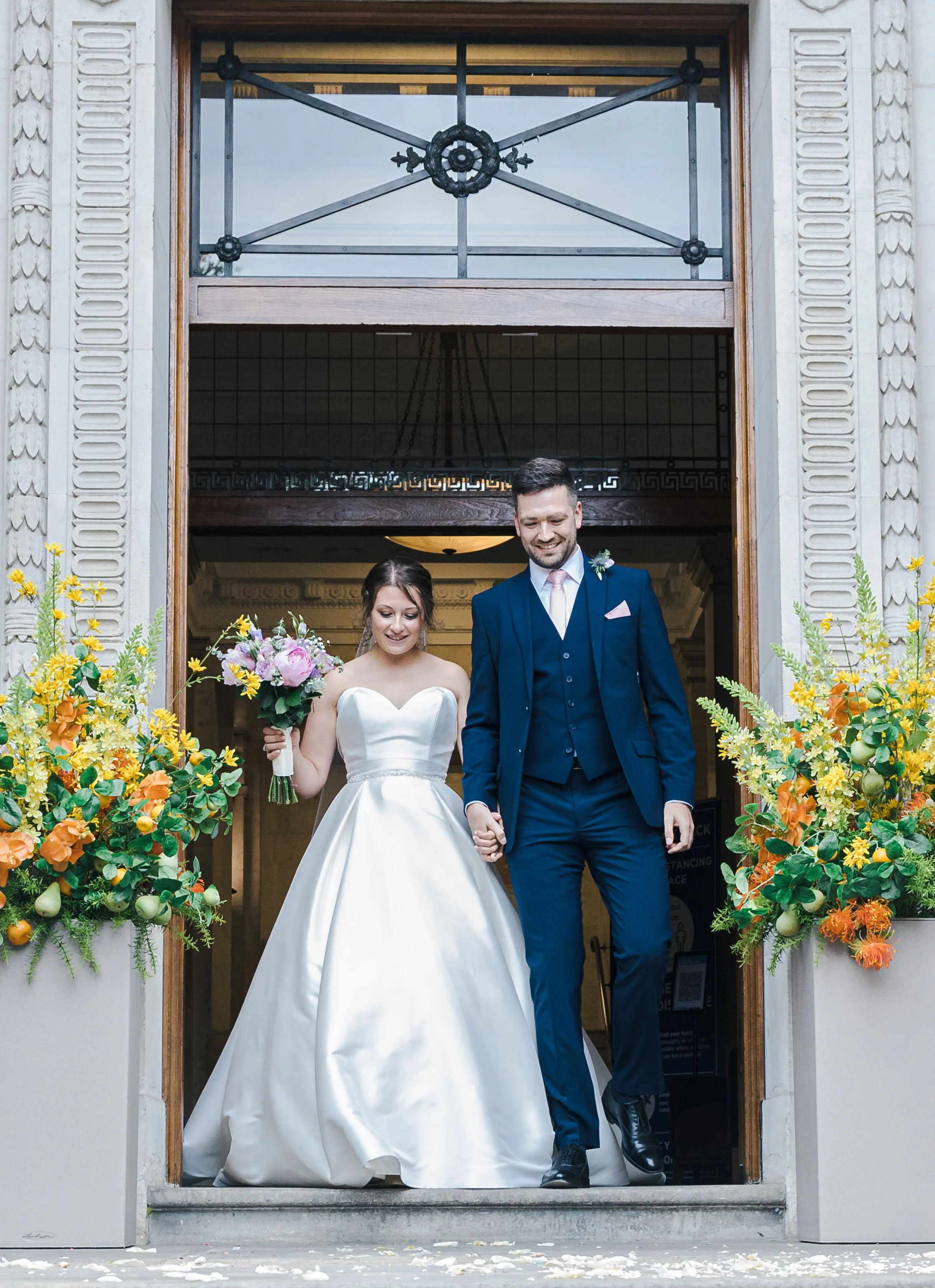 A bride and groom walking out of a building hand in hand, smiling. The bride is holding a bouquet of flowers, and both are dressed in wedding attire. There are large flower arrangements on either side of the doorway.