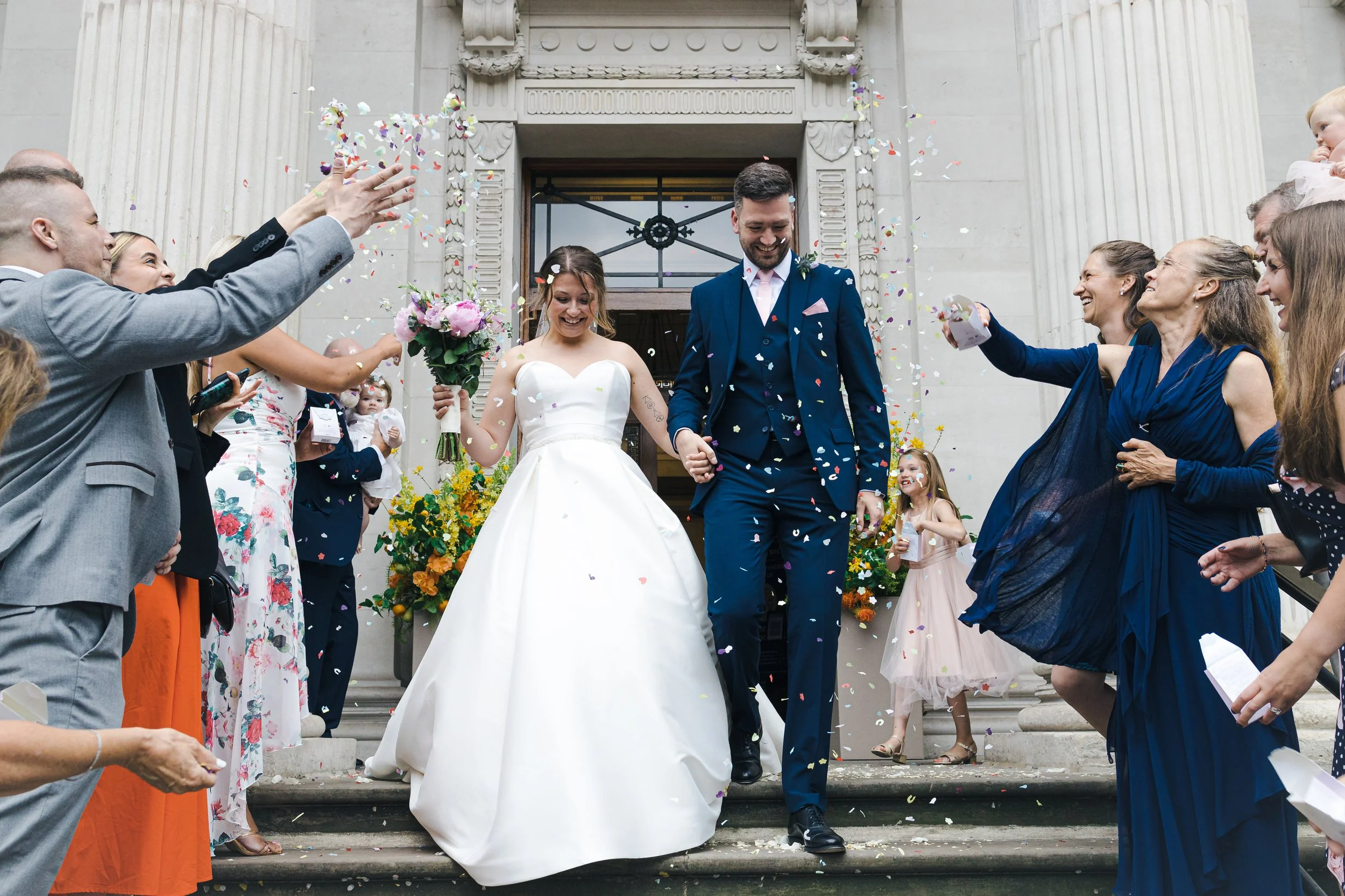 Bride and groom walking down steps outside a church, surrounded by friends and family throwing confetti.