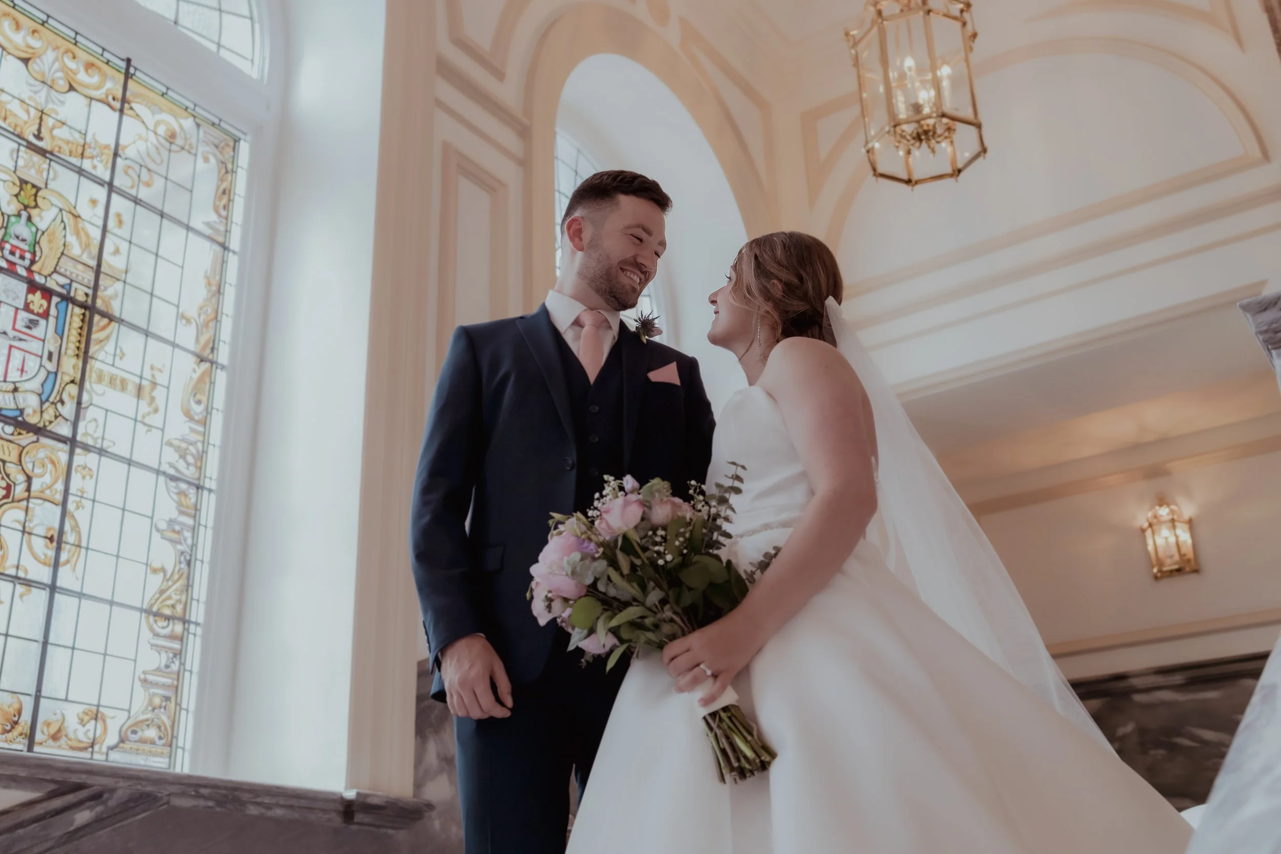 A bride and groom with big smiles on their faces, standing inside a wedding ceremony venue with stained glass and elegant lighting.
