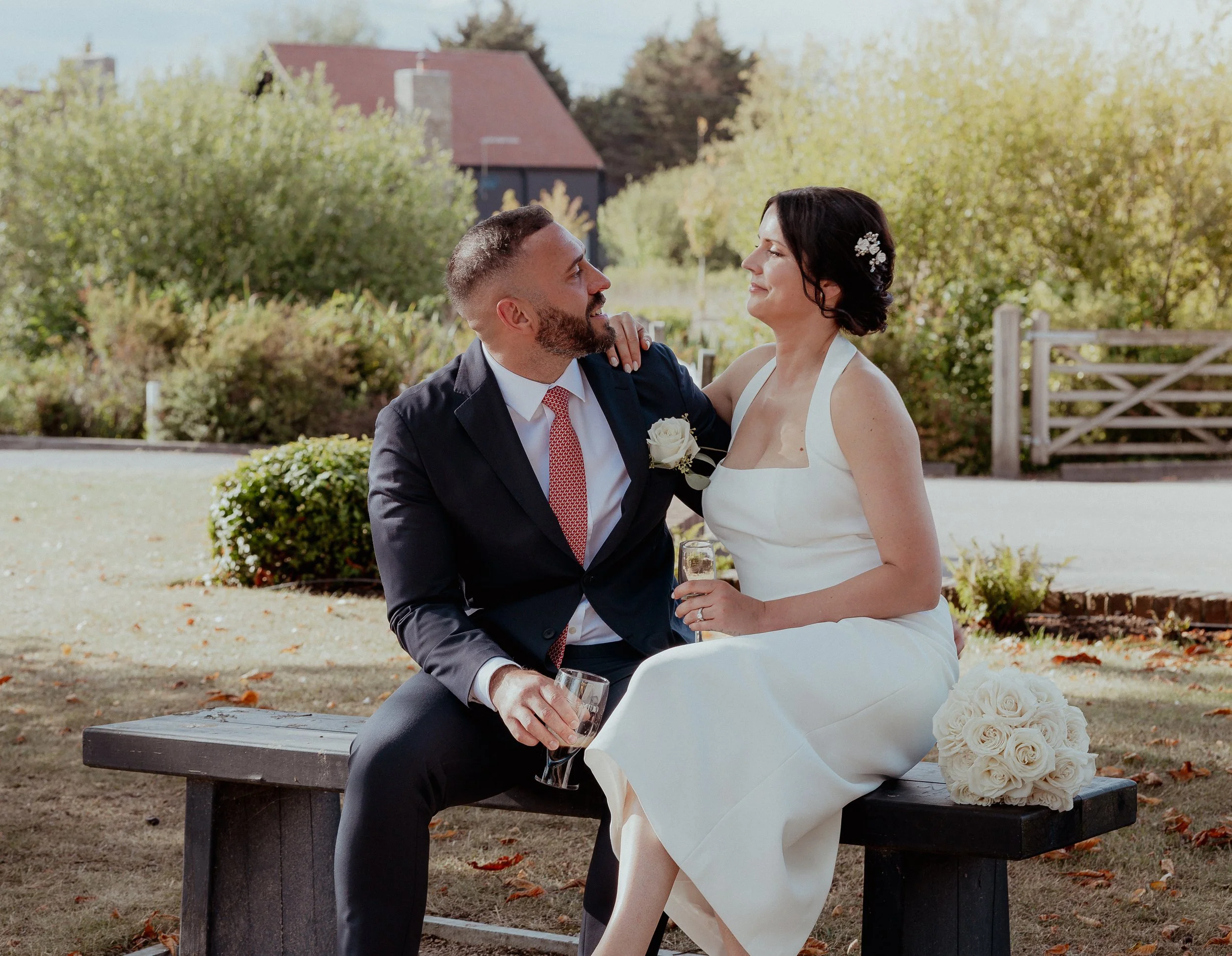 A couple dressed in wedding attire sitting on a wooden bench outdoors, smiling at each other, with a bouquet of white roses beside them and a scenic background of trees and a house.