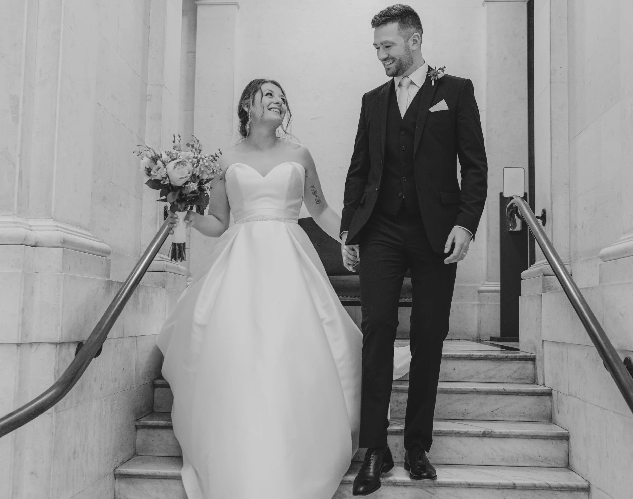 A bride and groom walking down a marble staircase, holding hands, smiling at each other during a wedding ceremony. The bride wears a strapless wedding gown and holds a bouquet, while the groom is dressed in a dark suit with a boutonniere.