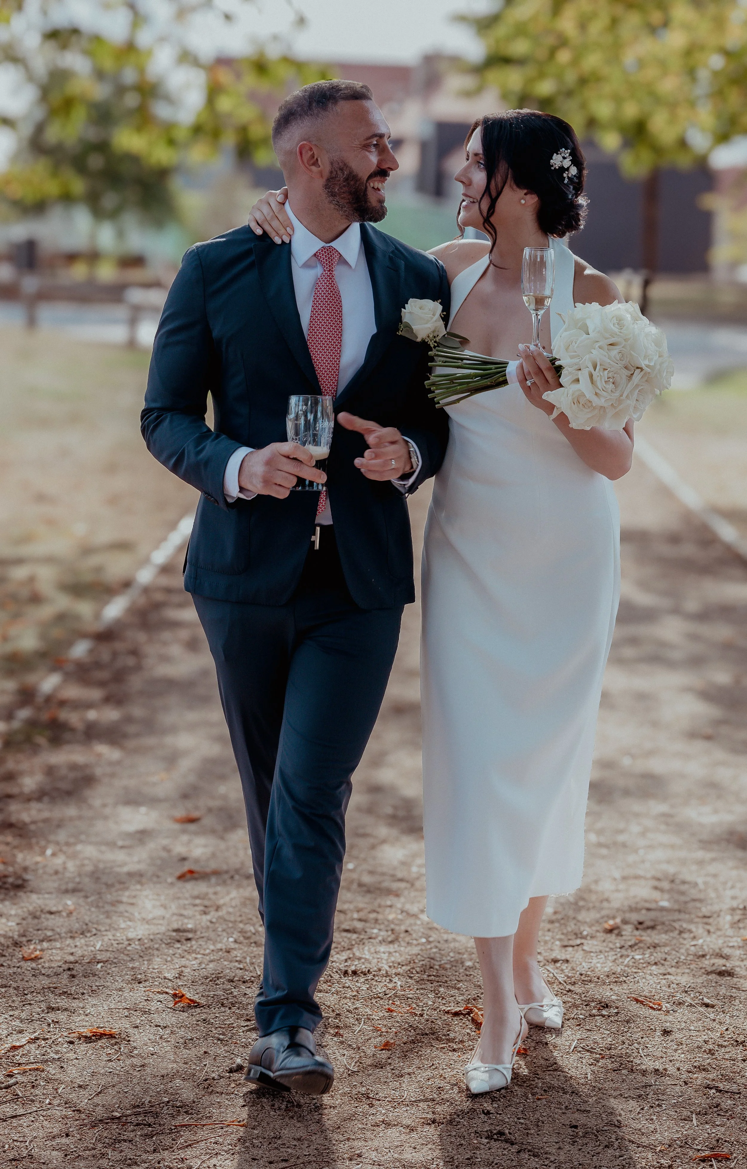 A newlywed couple walking outdoors, the bride in a white gown holding a bouquet of white roses and a champagne glass, the groom in a dark suit holding a glass of champagne, smiling at each other.