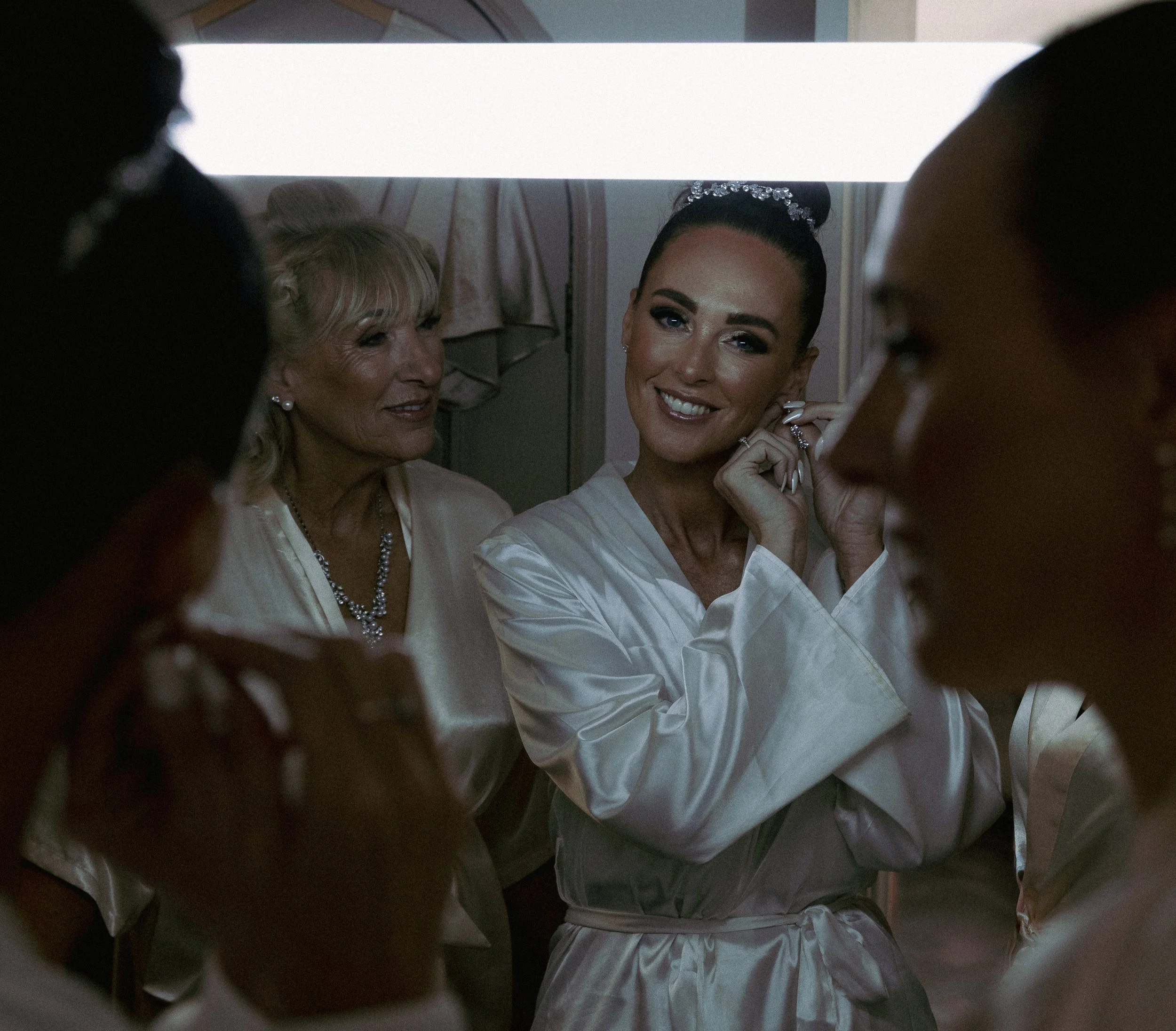 A smiling bride with dark hair styled in an updo, wearing a satin robe and earrings, surrounded by women in robes, preparing for her wedding.