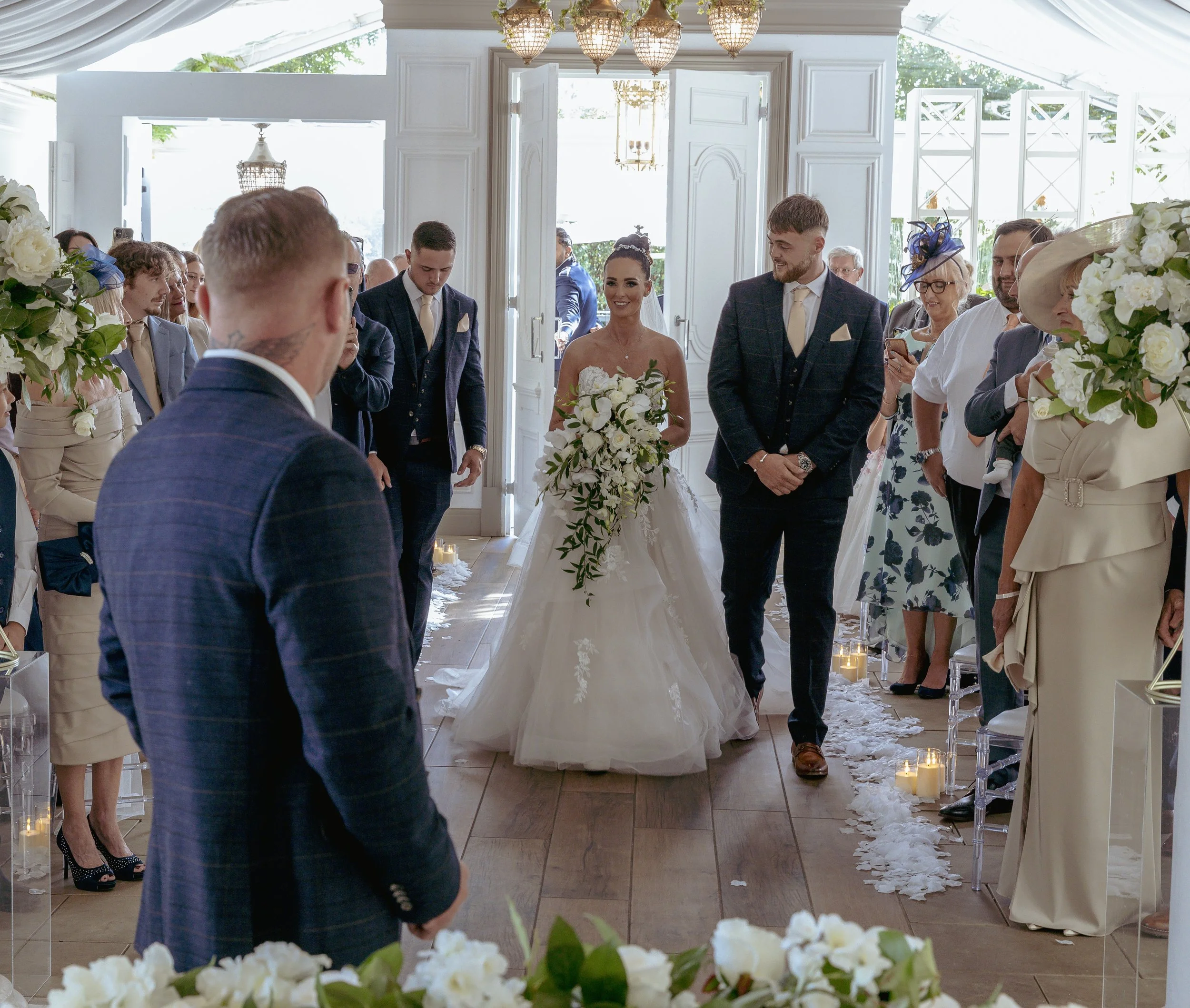 A bride and groom walking down the aisle at their wedding ceremony, surrounded by guests in a bright, elegant venue decorated with flowers and candles.