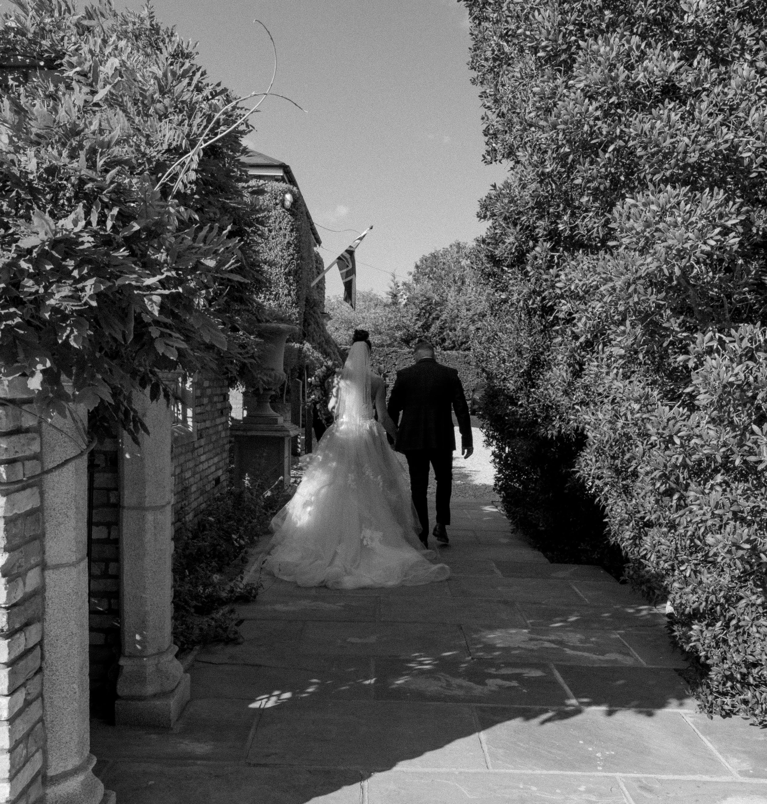 A bride and groom walking hand in hand on a garden pathway on their wedding day, surrounded by trees and foliage.