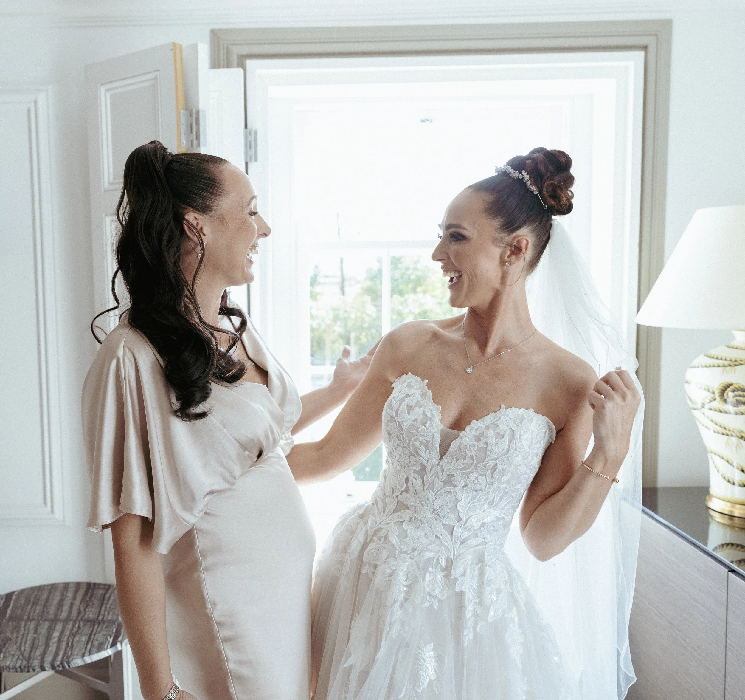 Two women, one in a wedding dress and the other in a light-colored dress, smiling and celebrating inside a room near a window.