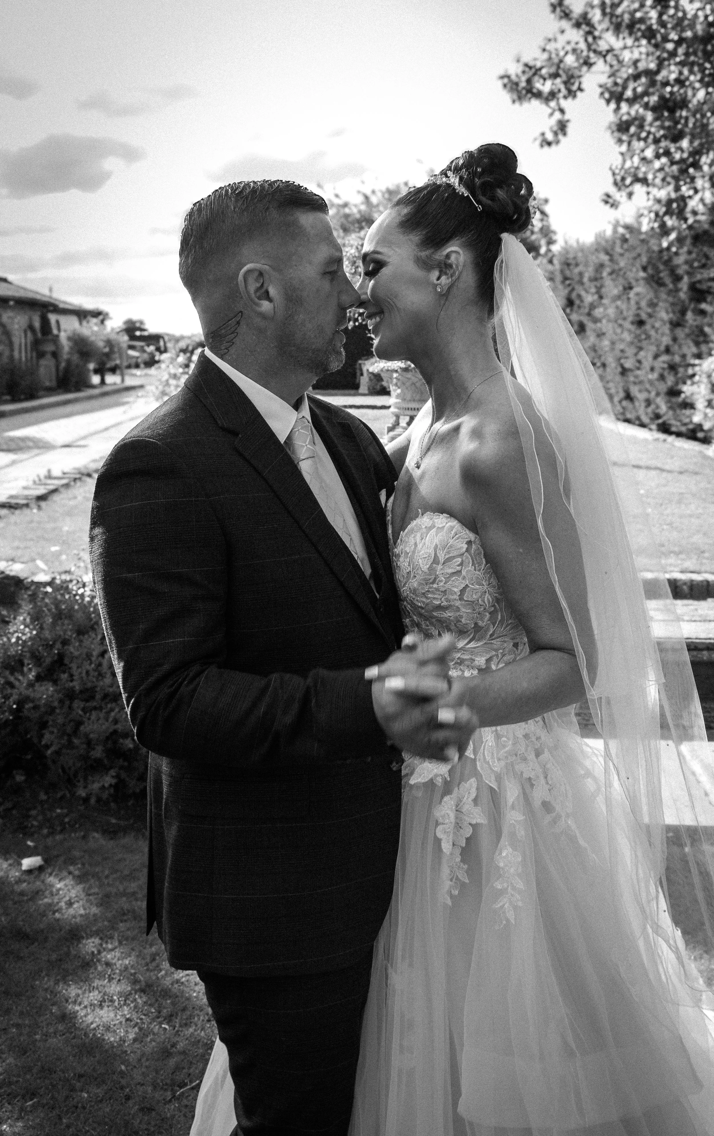 A black-and-white photo of a bride and groom standing close together, smiling and about to kiss outdoors on their wedding day. The bride is wearing a strapless lace wedding gown with a veil, and the groom is dressed in a suit with a tattoo on his nec