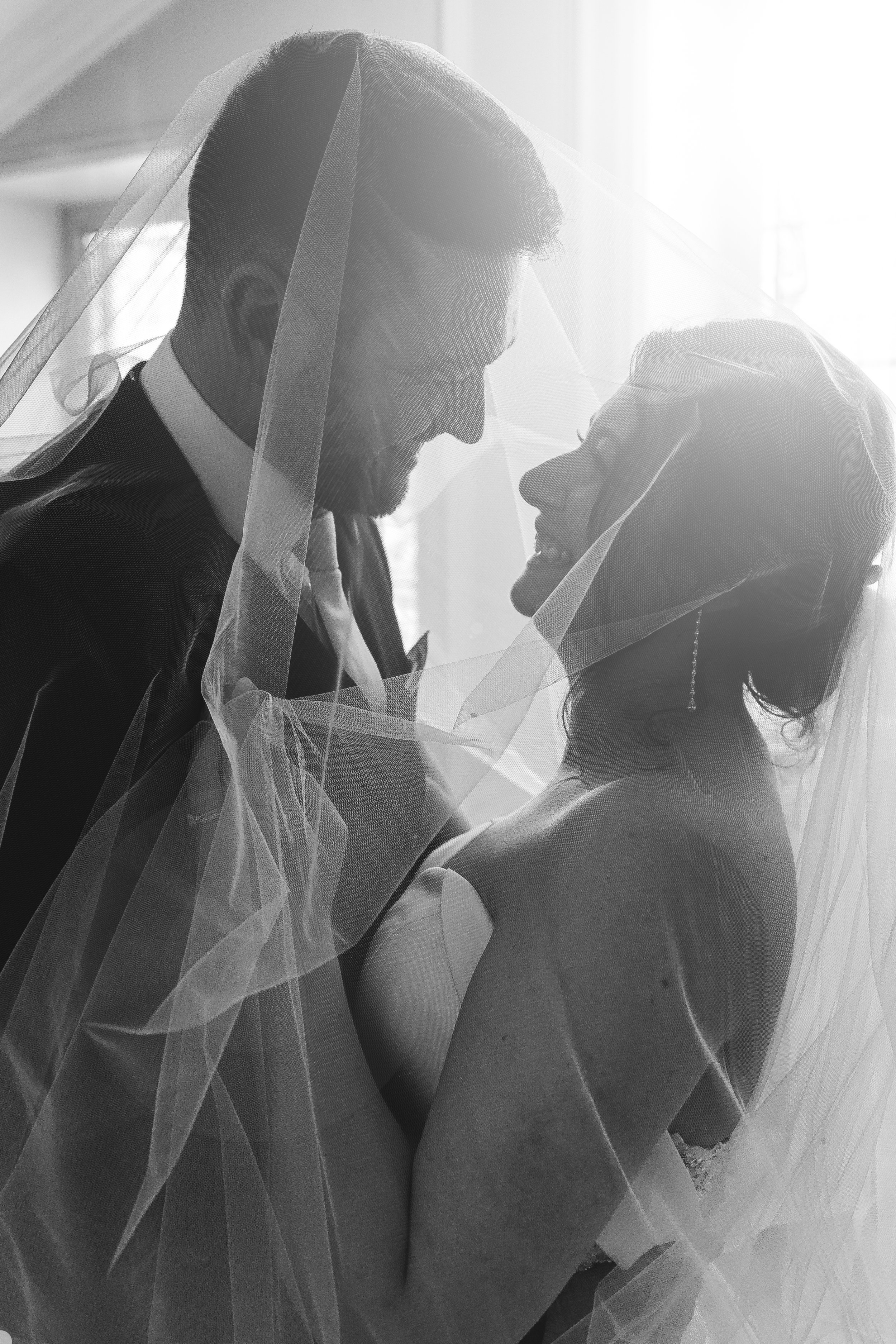 Black and white photo of a bride and groom under a veil, looking at each other, smiling, in a wedding setting.